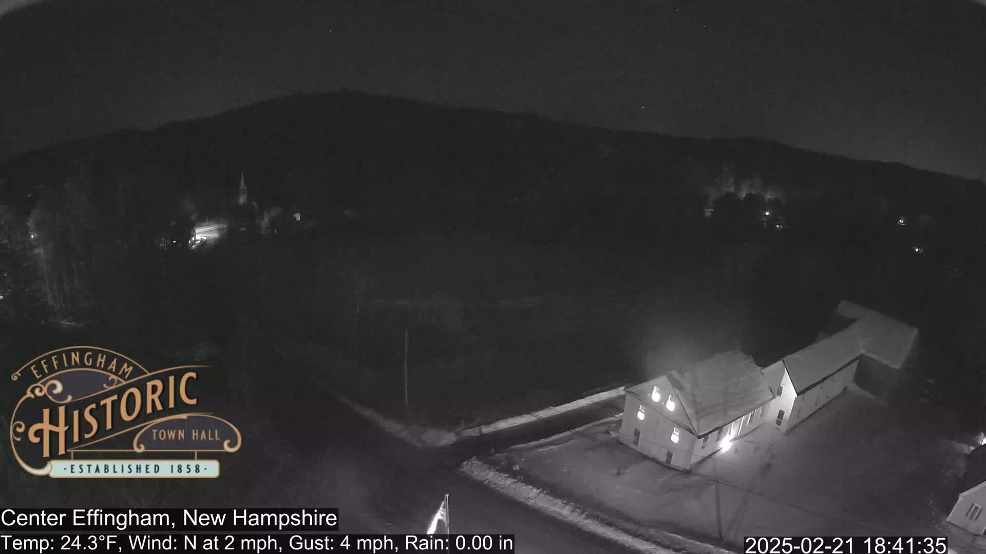 A nighttime, low-light view of a small town with light rain and a mostly clear sky shows a building with lights on and a few other lights scattered throughout the town.