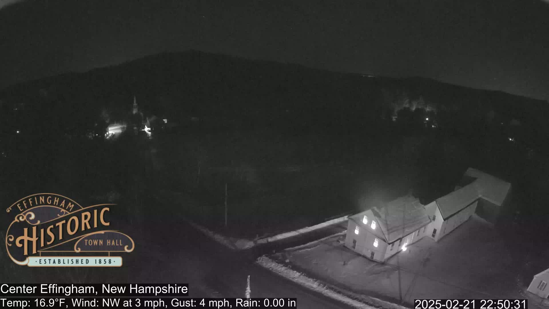A nighttime, low-light aerial view of a small town shows a building illuminated and a light dusting of snow on the ground, with a northwest wind at 3 mph and gusts up to 4 mph.