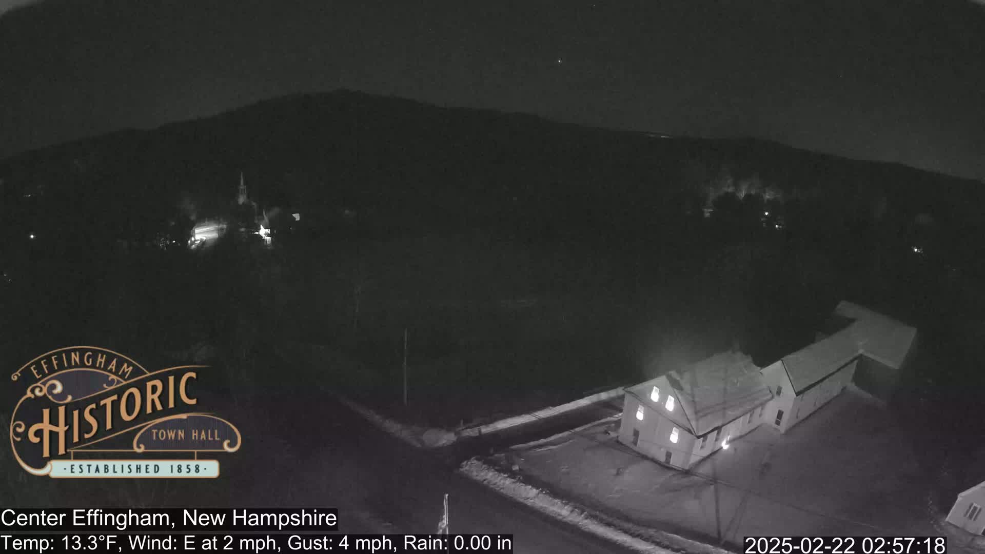 A nighttime, aerial view of a small town with a building illuminated, under a clear sky with light snowfall on the ground.