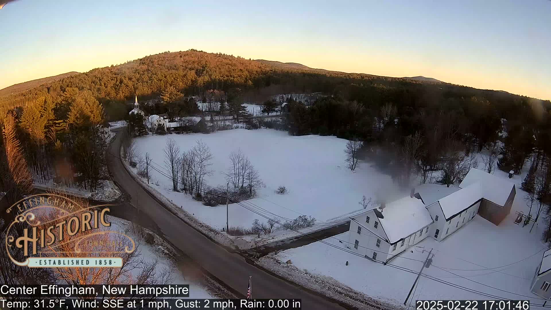 A snow-covered rural landscape with a road, buildings, and a church steeple is visible under a clear, twilight sky.