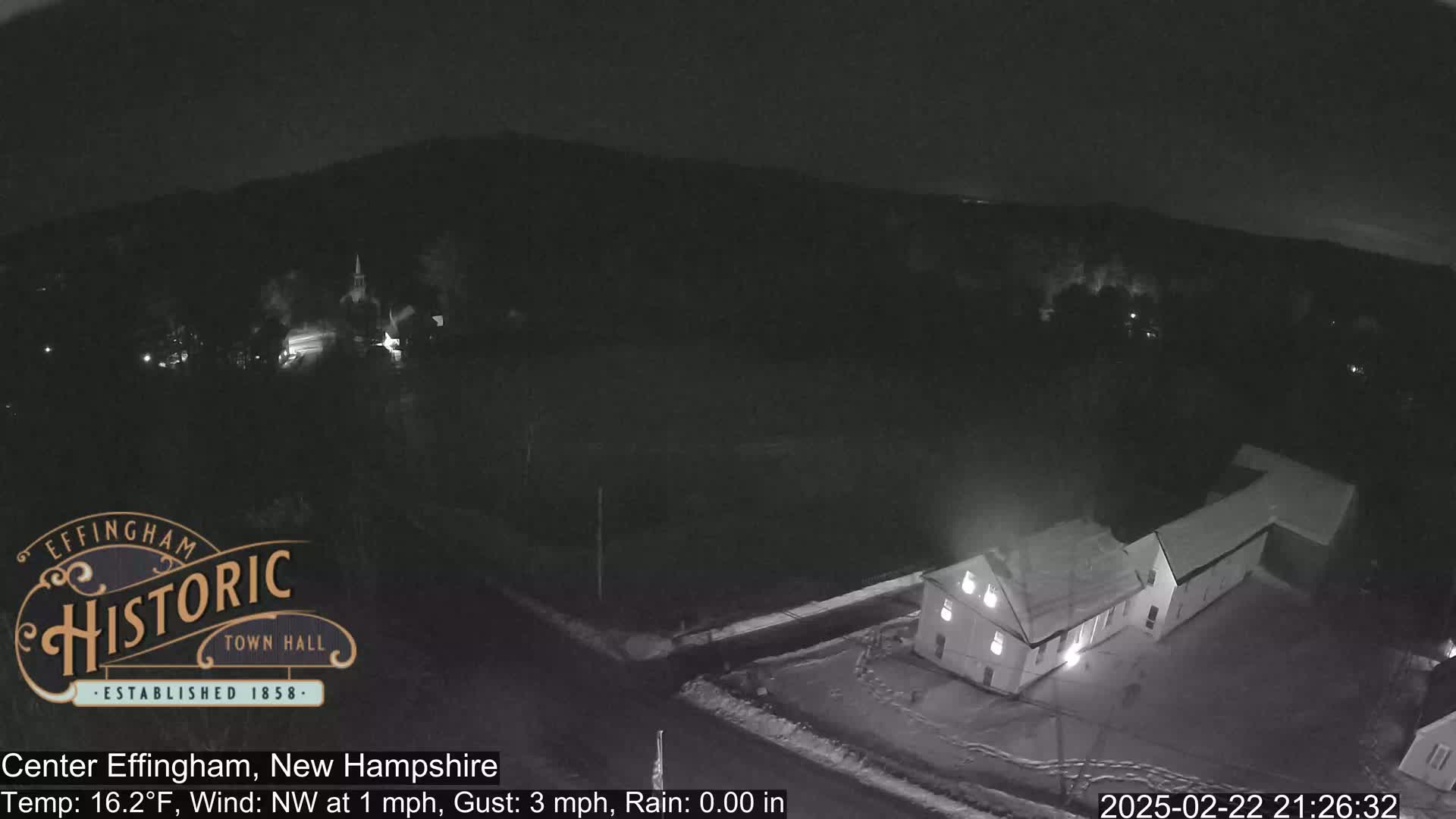 A nighttime, snowy scene shows a building and a town with a church steeple under a dark sky with a light northwest wind.