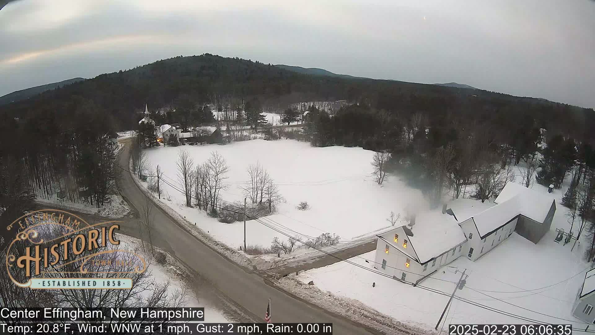 A snow-covered road leads through a small, snow-covered town nestled amongst snow-covered hills and trees under an overcast sky.