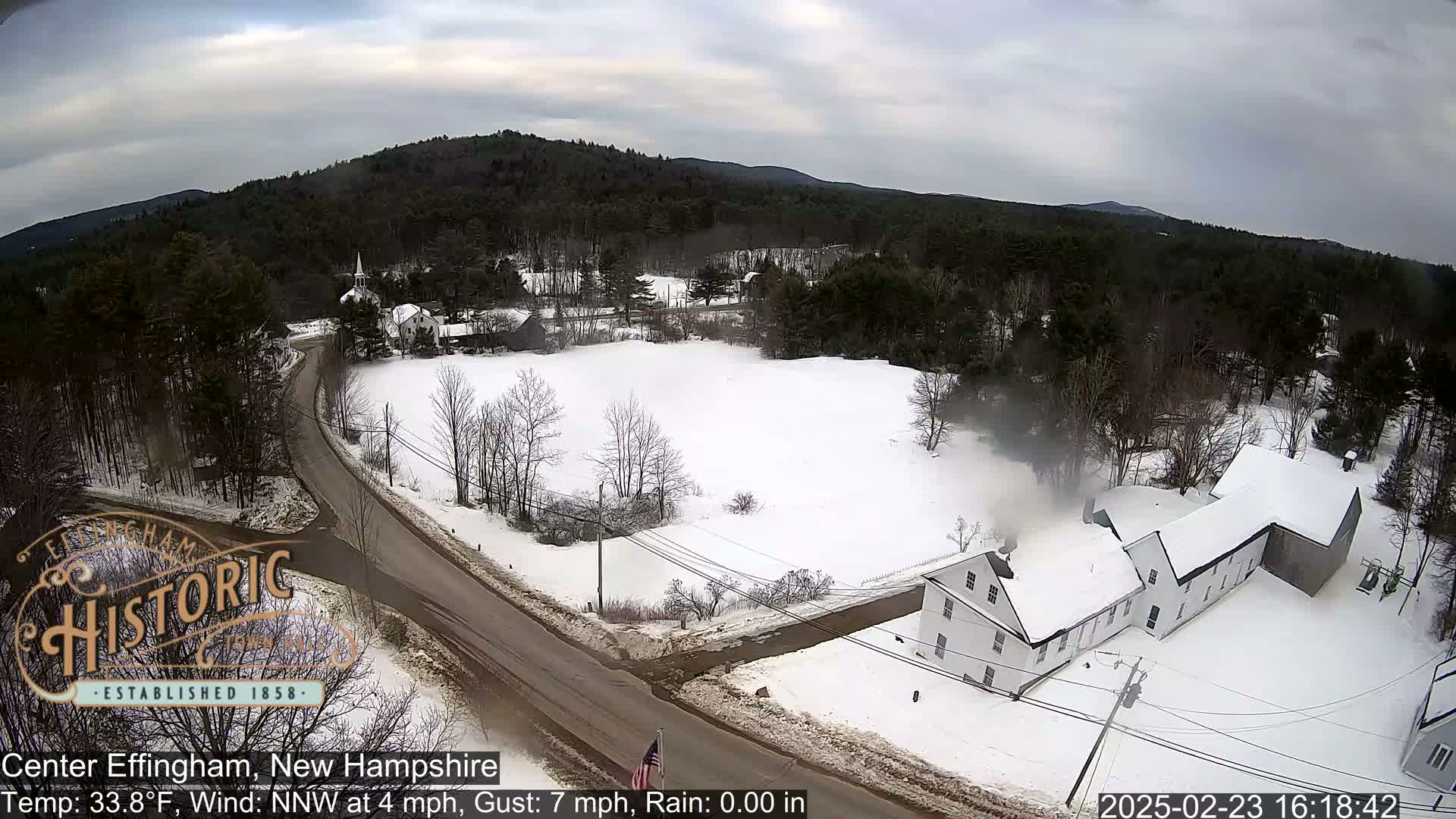A snow-covered landscape with a road, buildings, and trees under a cloudy sky.