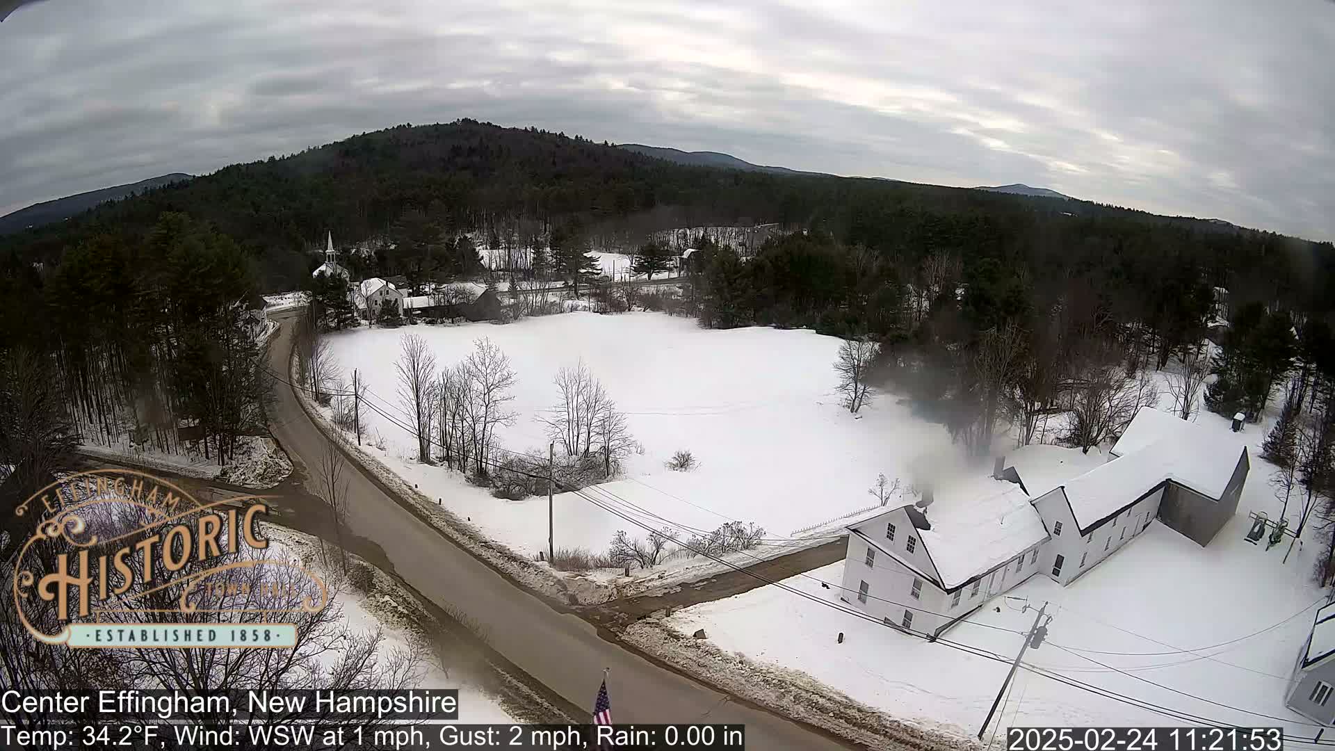 A snow-covered rural landscape with a long white building near a road, under an overcast sky.