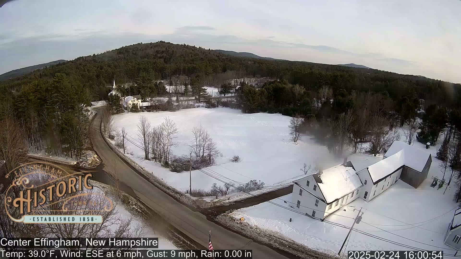A snow-covered rural landscape with a road, buildings, and a forest in the background under a partly cloudy sky.