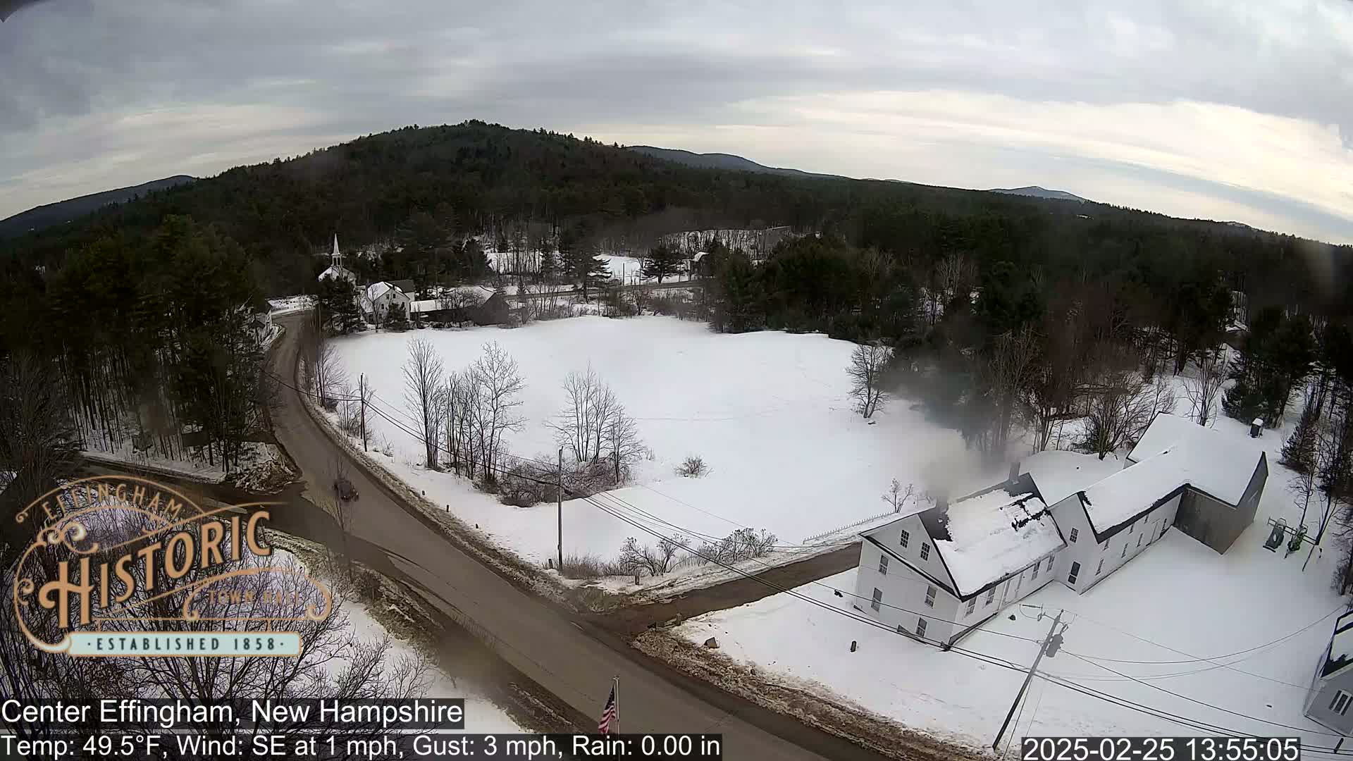 A snow-covered rural scene with a road, buildings, and trees under an overcast sky.