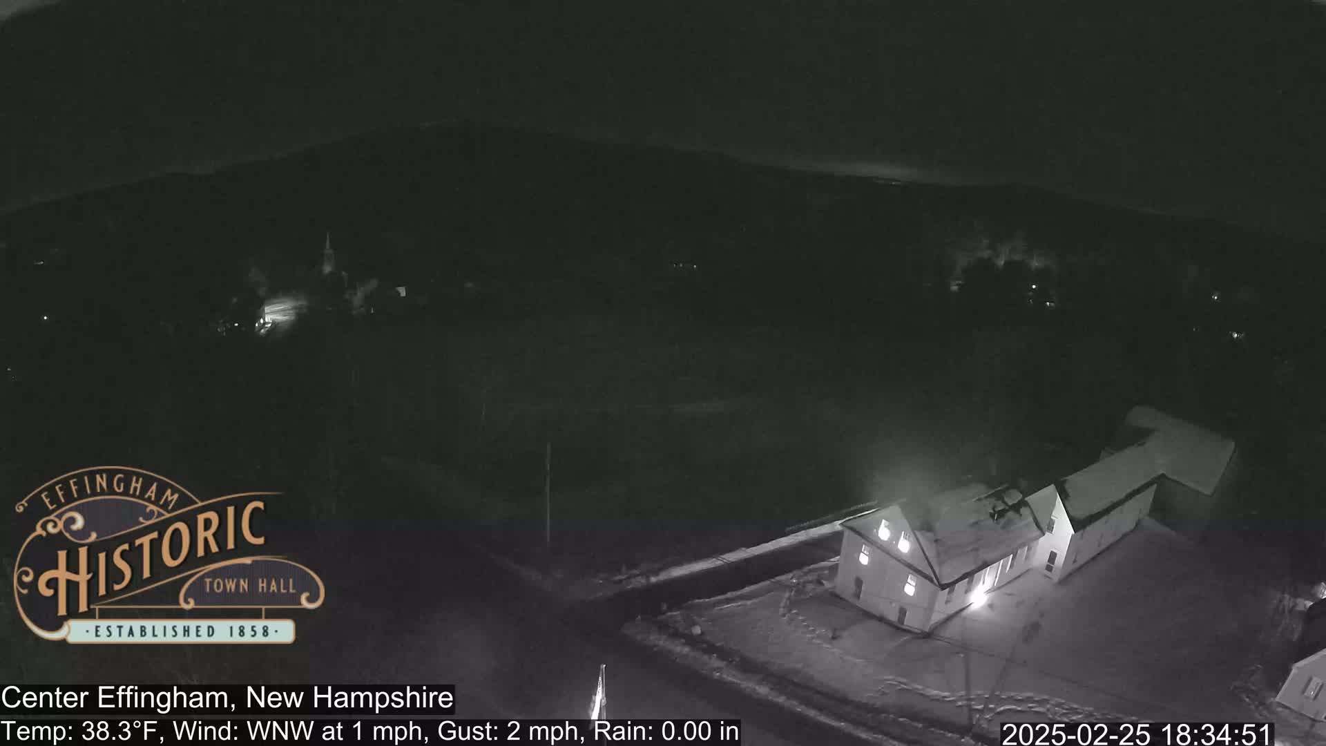 A nighttime, snow-covered landscape is seen from an elevated perspective, showing a building illuminated near a road, with a calm wind and no rain.