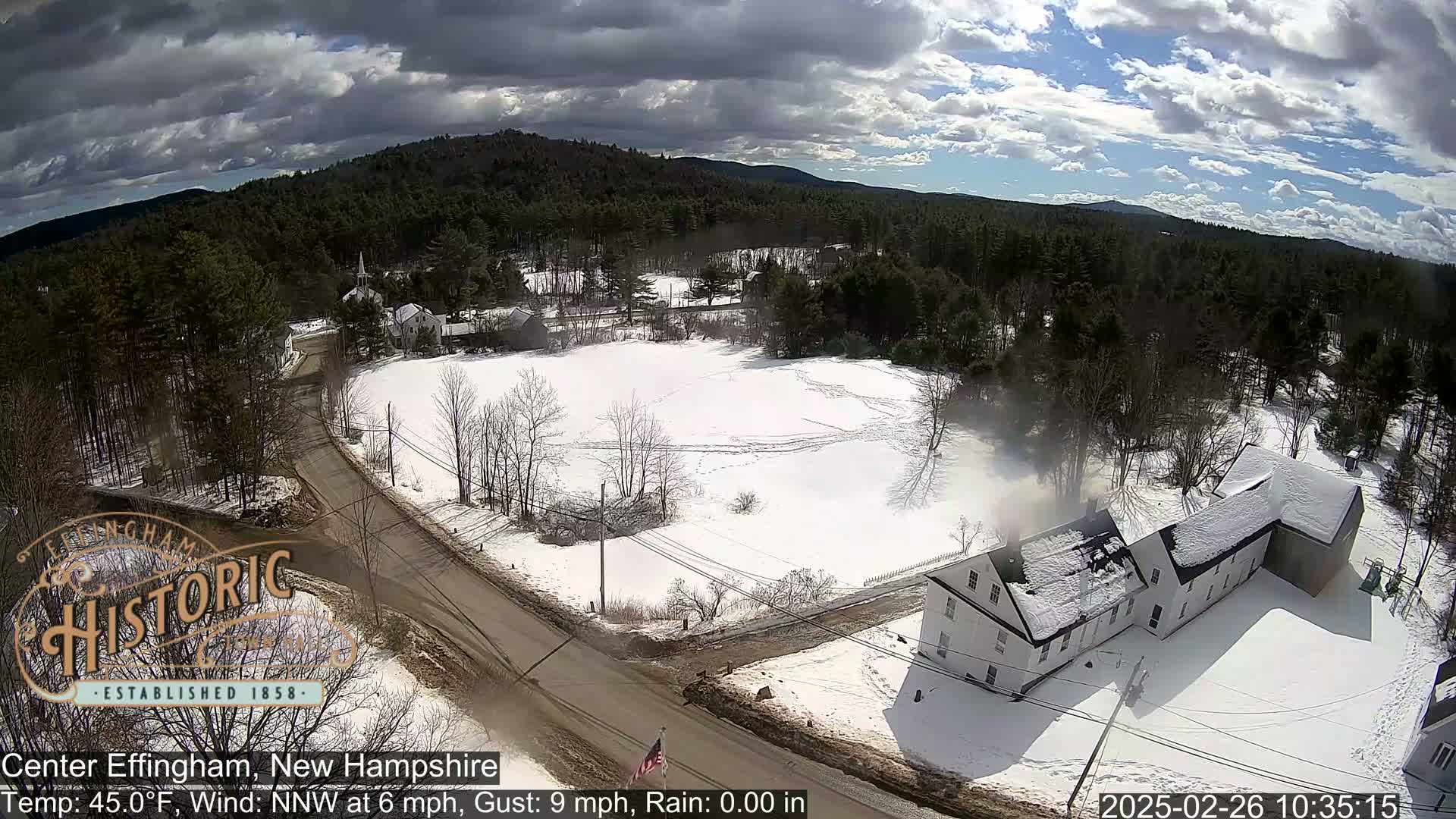A snow-covered rural landscape with a road, houses, and a forest under a partly cloudy sky.