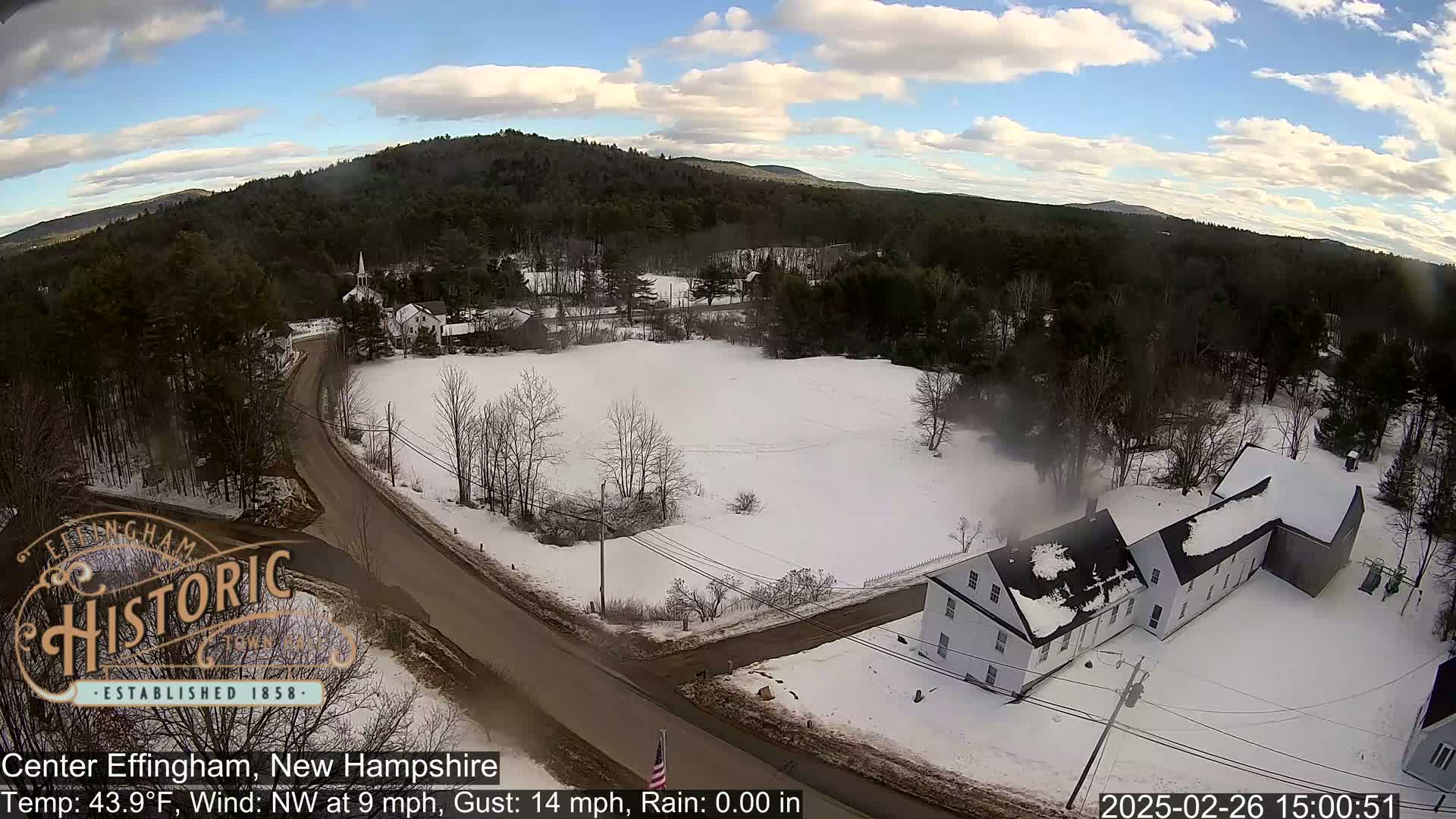 A snow-covered rural landscape with a road, buildings, and trees under a partly cloudy sky.
