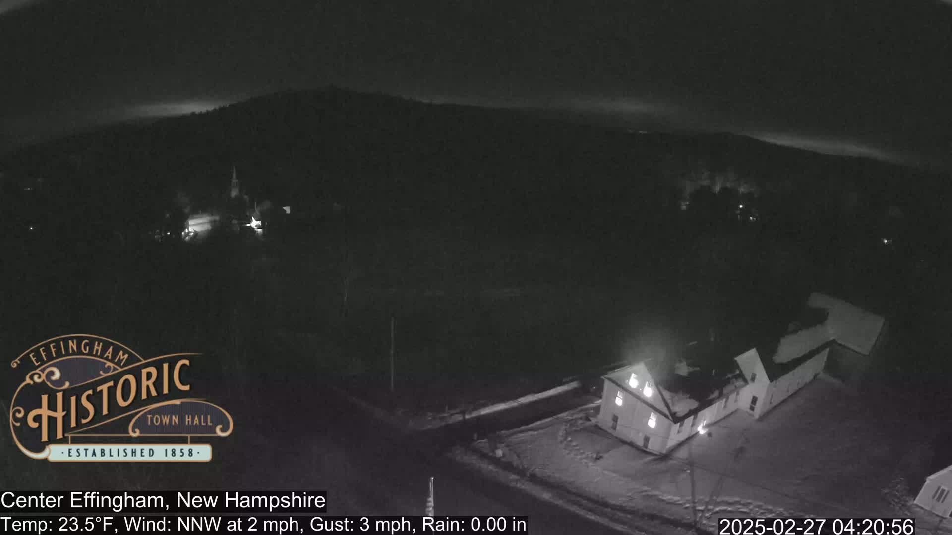 A nighttime, snow-covered landscape shows a building with lights on and a hilly, dark background under a clear sky.