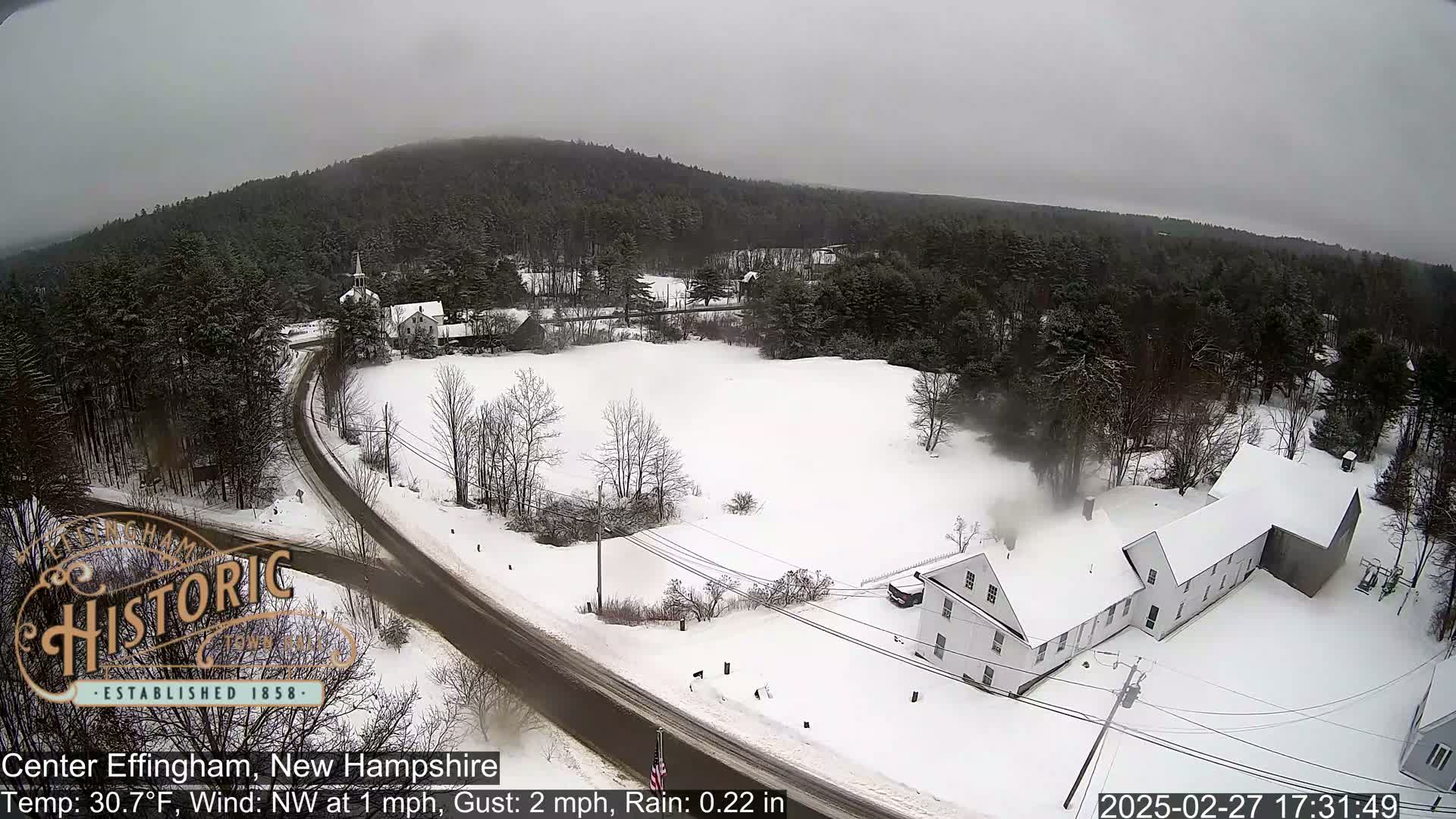 A snow-covered rural landscape with a road, buildings, and trees under an overcast sky.