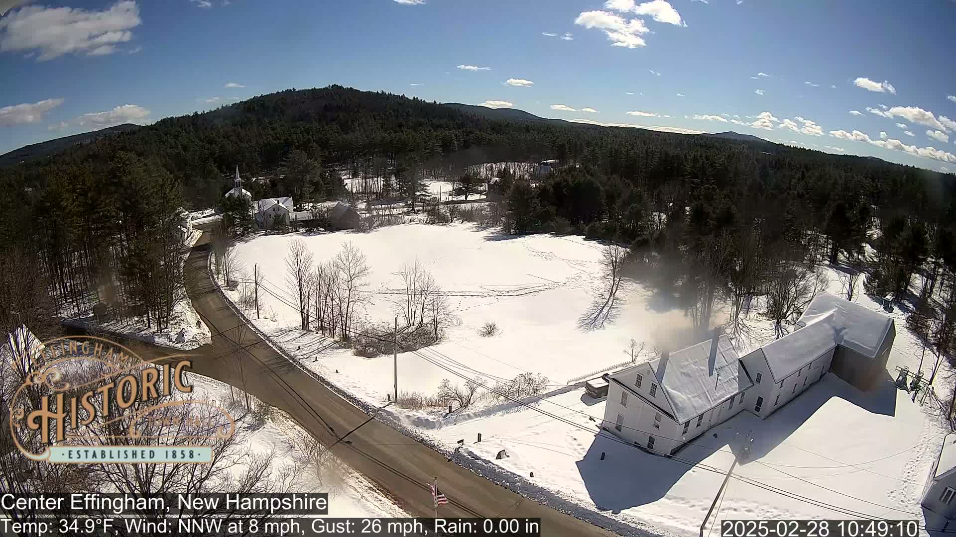 A snow-covered rural scene with a road, buildings, and a background of dark-green wooded hills under a mostly sunny sky.