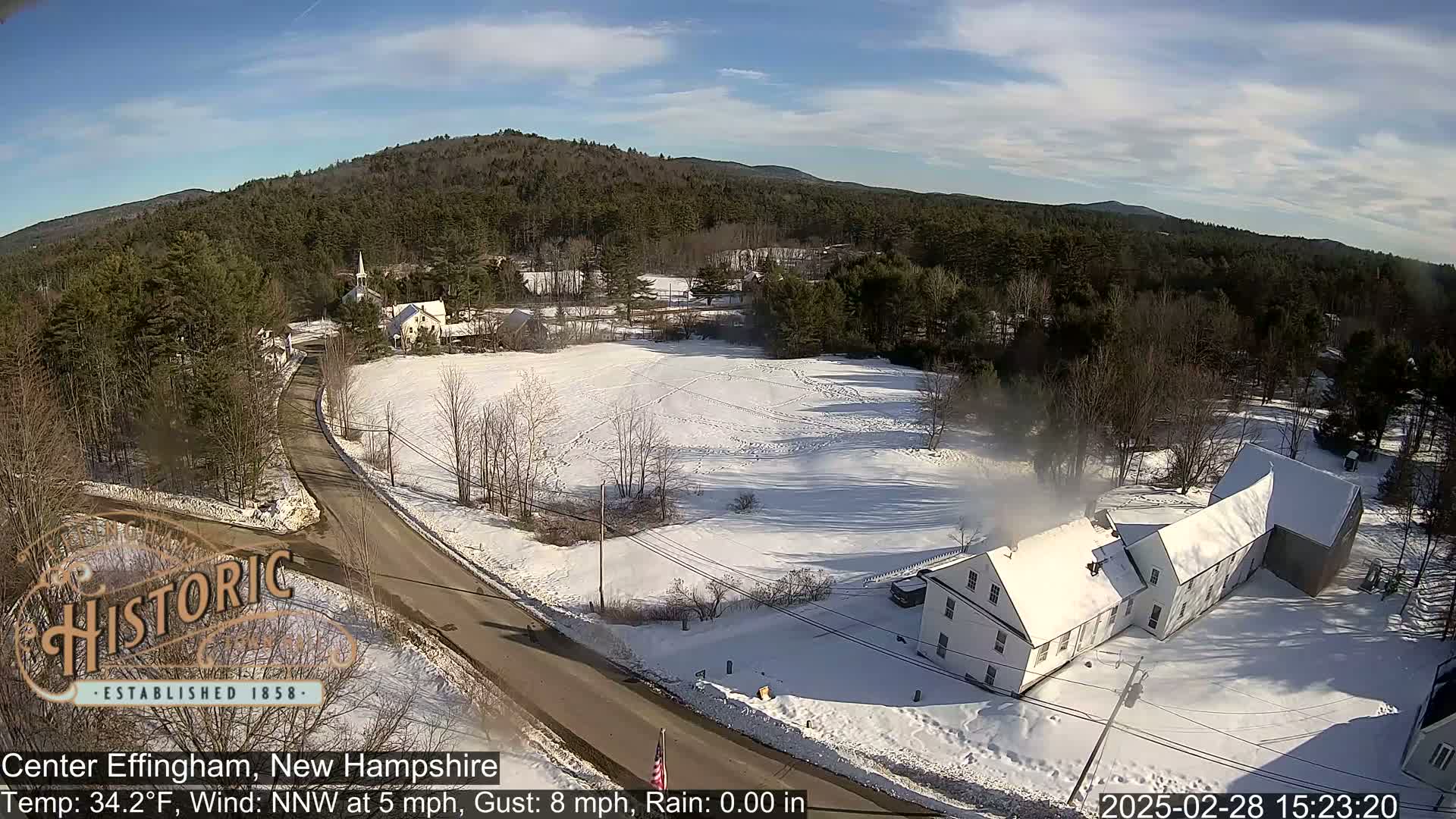 A snow-covered rural landscape with a road, buildings, and trees under a mostly sunny sky.