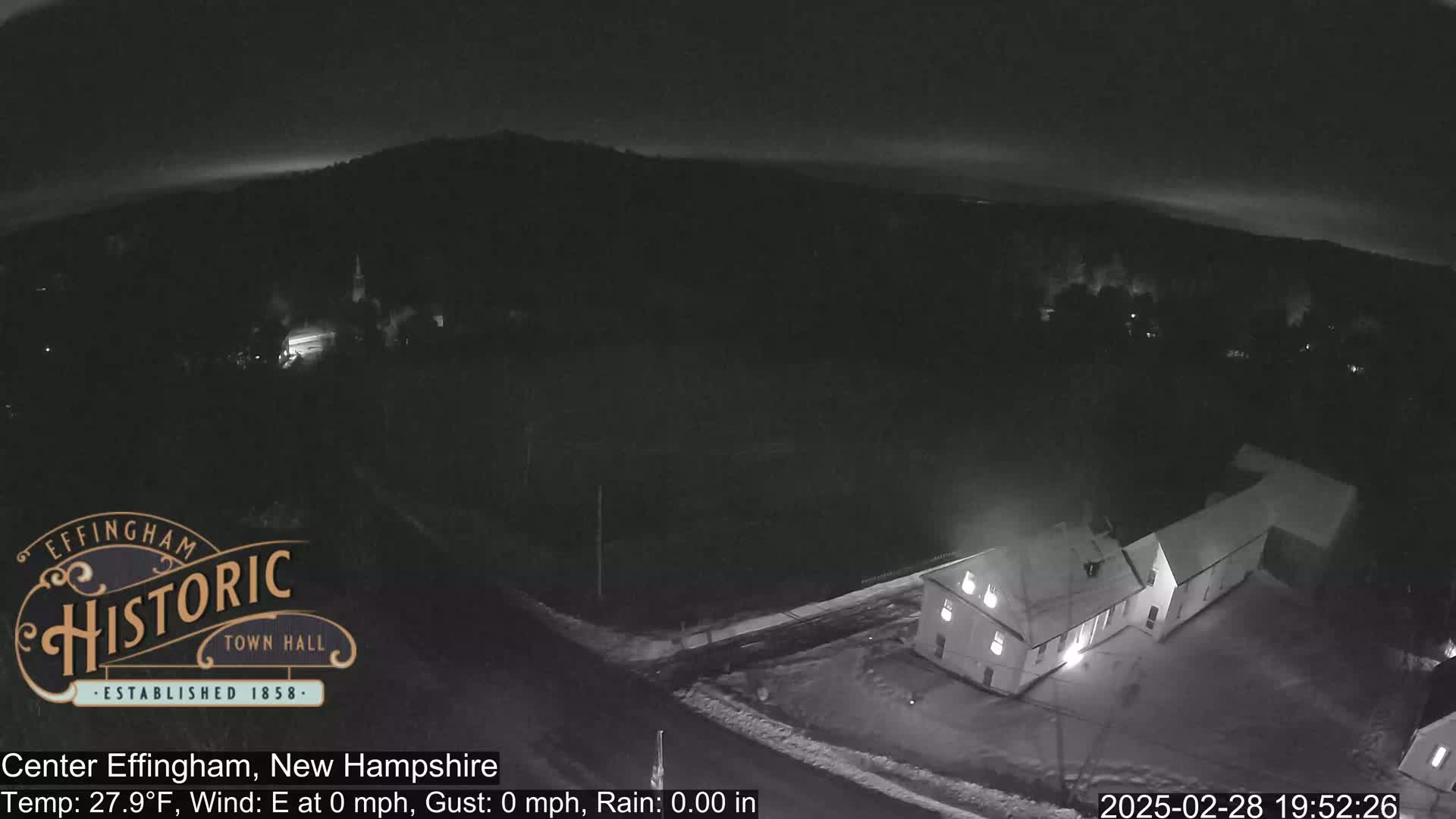 A nighttime, rainy scene shows a building illuminated, with snow on the ground and a dark, hilly landscape in the background.