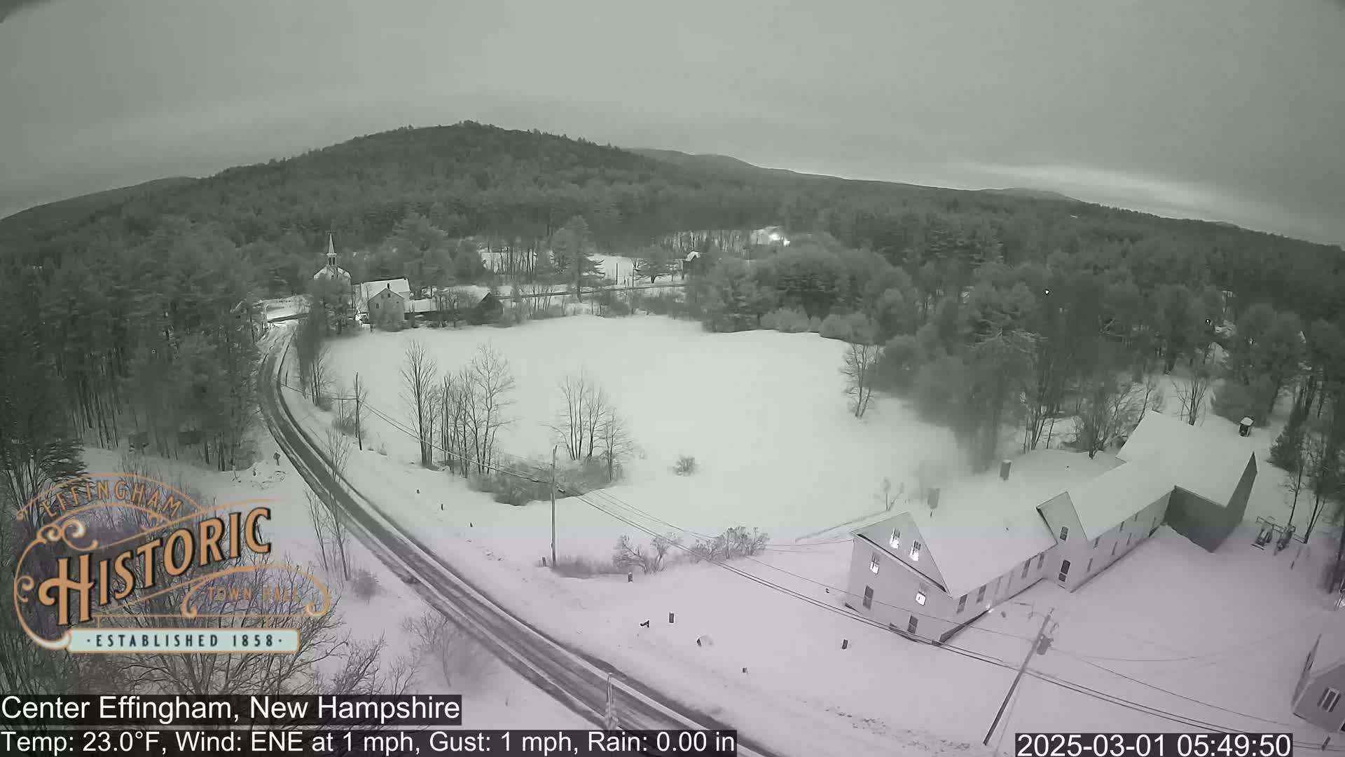 A snow-covered rural landscape with buildings, a road, and trees under an overcast sky.