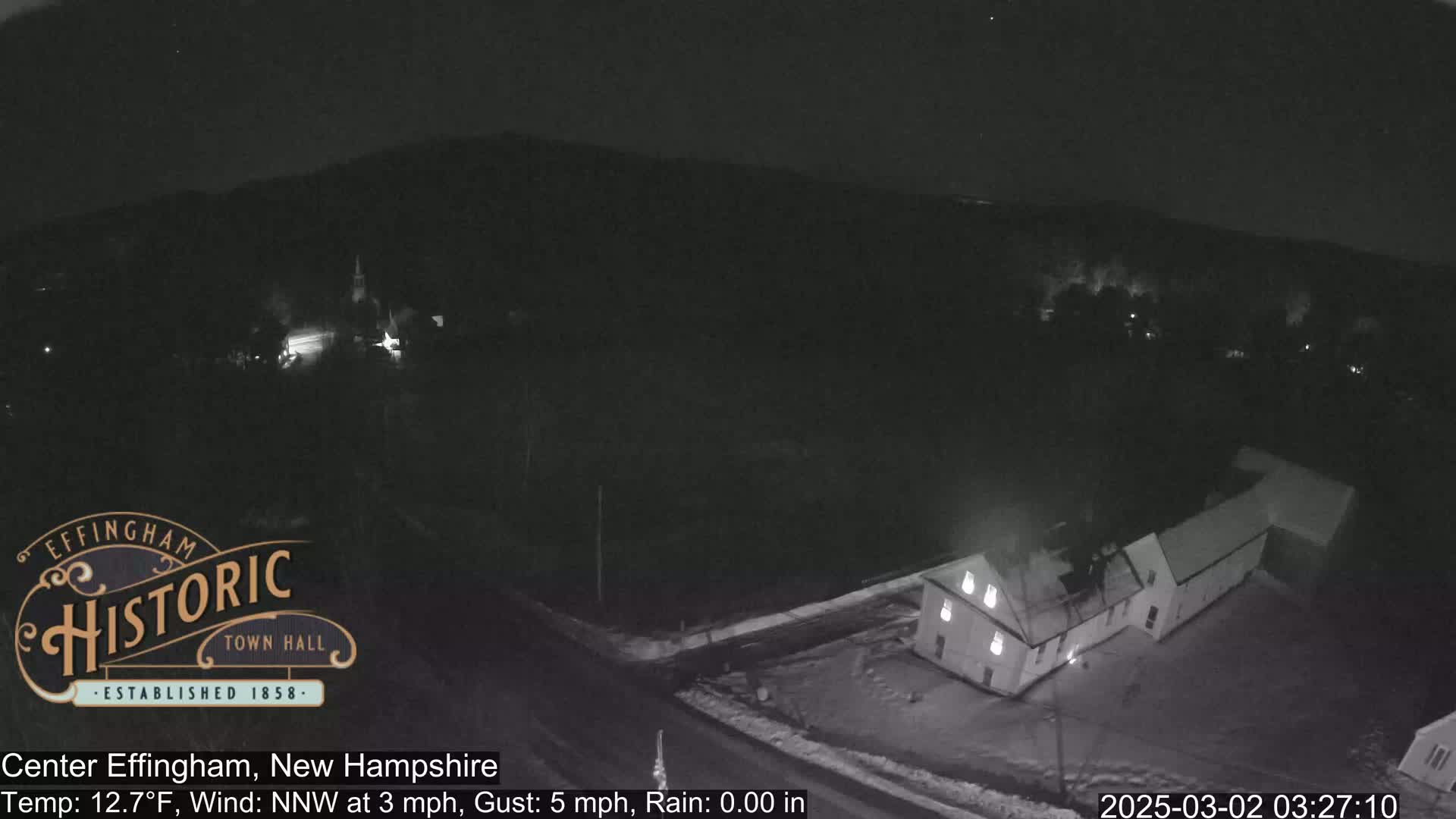 A nighttime, low-light aerial view of a small town with a building illuminated, under a clear sky, with a temperature of 12.7°F and a north-northwest wind.