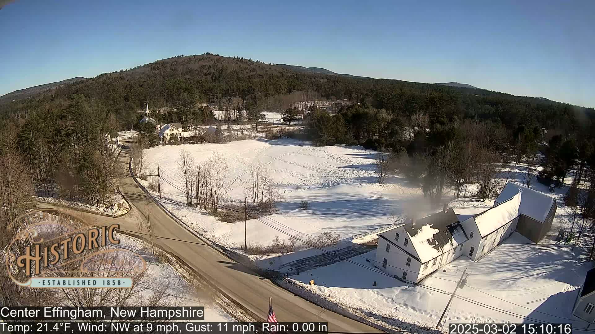 A snow-covered rural area with a road, buildings, and trees under a clear, sunny sky.