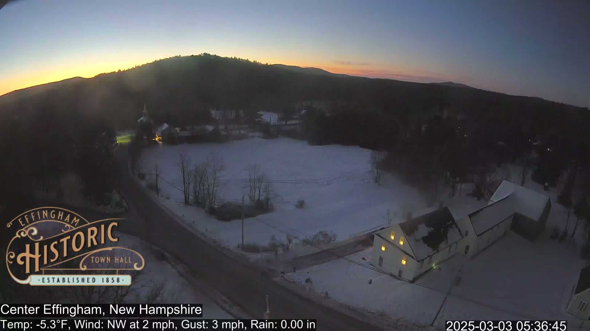 A snow-covered landscape at sunrise, showing a building with lit windows, a road, and a hill in the background.