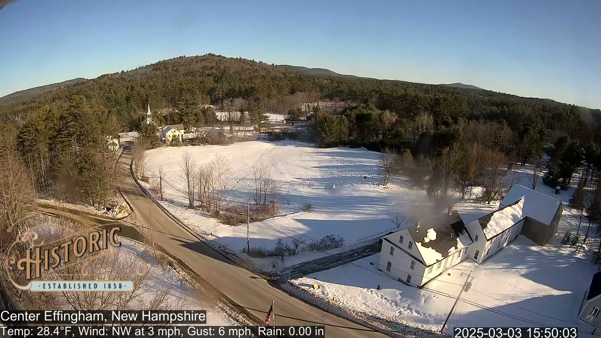 A snow-covered village scene with a road, buildings, and a hilly, wooded background under a clear, sunny sky.