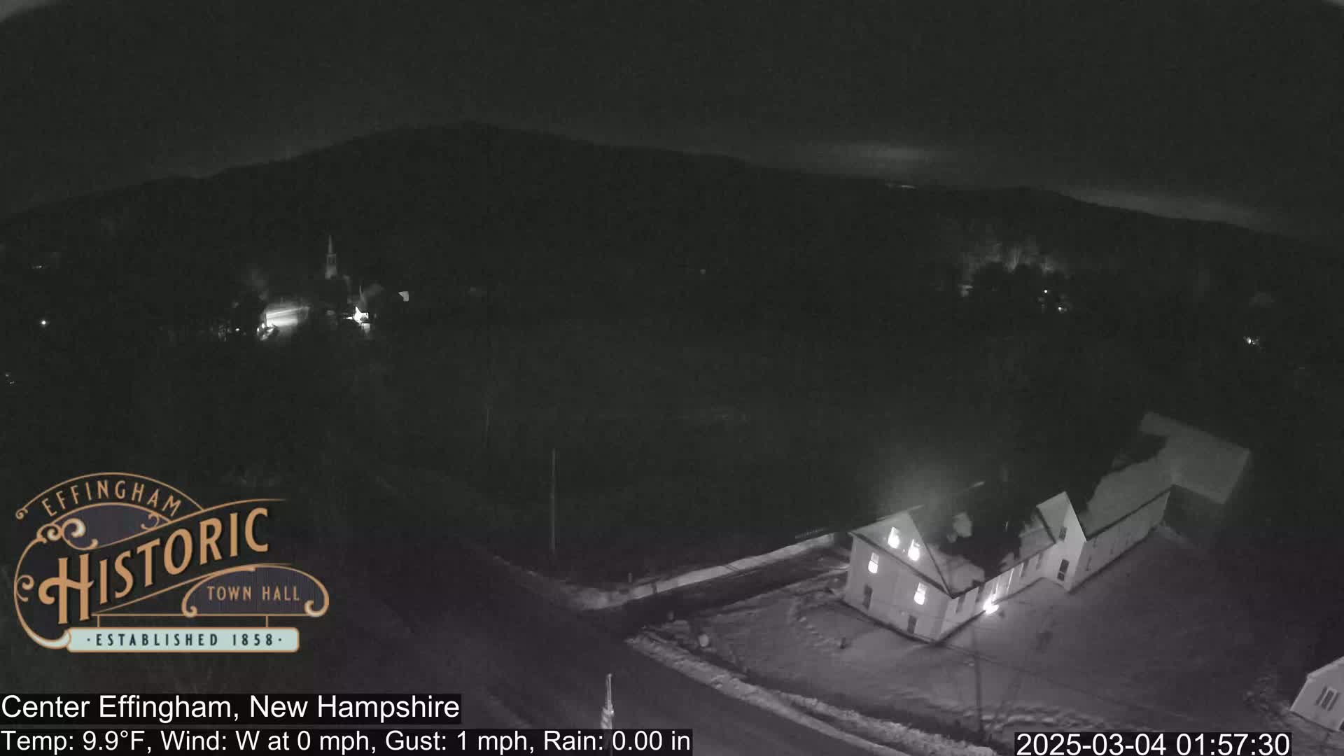 A nighttime, snow-covered scene shows a small town with a few lit buildings under a dark sky.