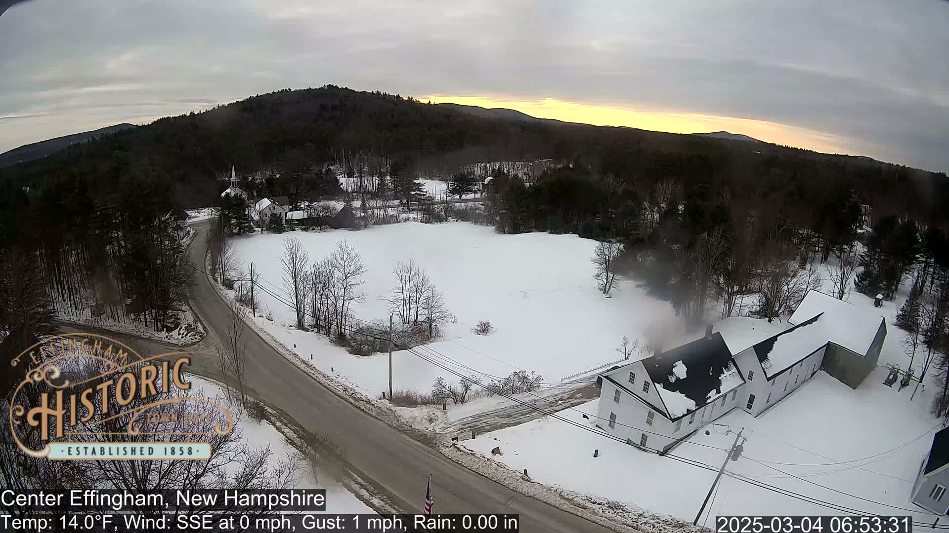 A snow-covered landscape at sunrise, featuring a road intersection, several buildings, and a wooded area in the background.