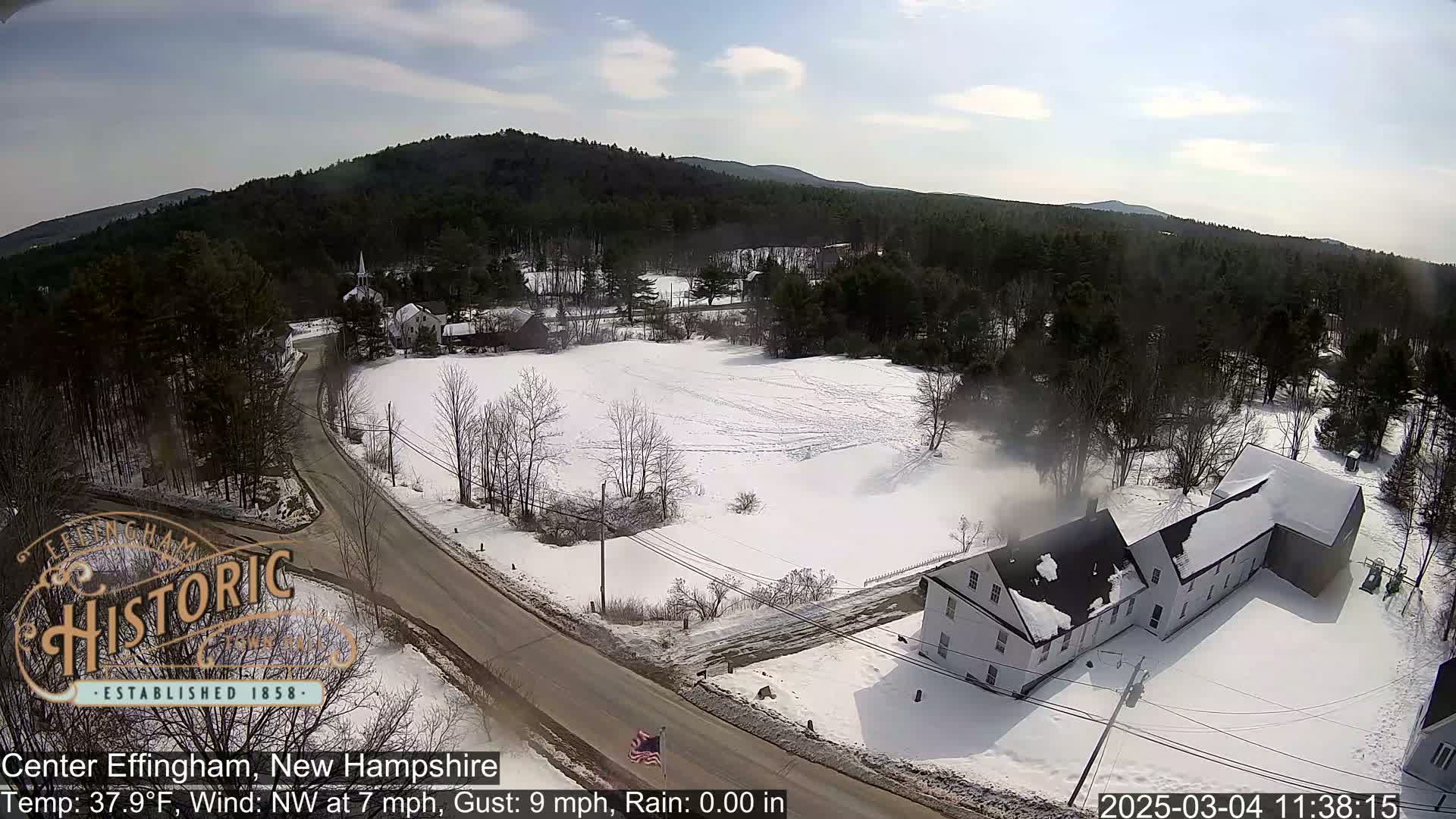 A snowy, partly sunny scene shows a road curving through a small town nestled among snow-covered fields and dark evergreen trees, with a long, white building prominent in the foreground.