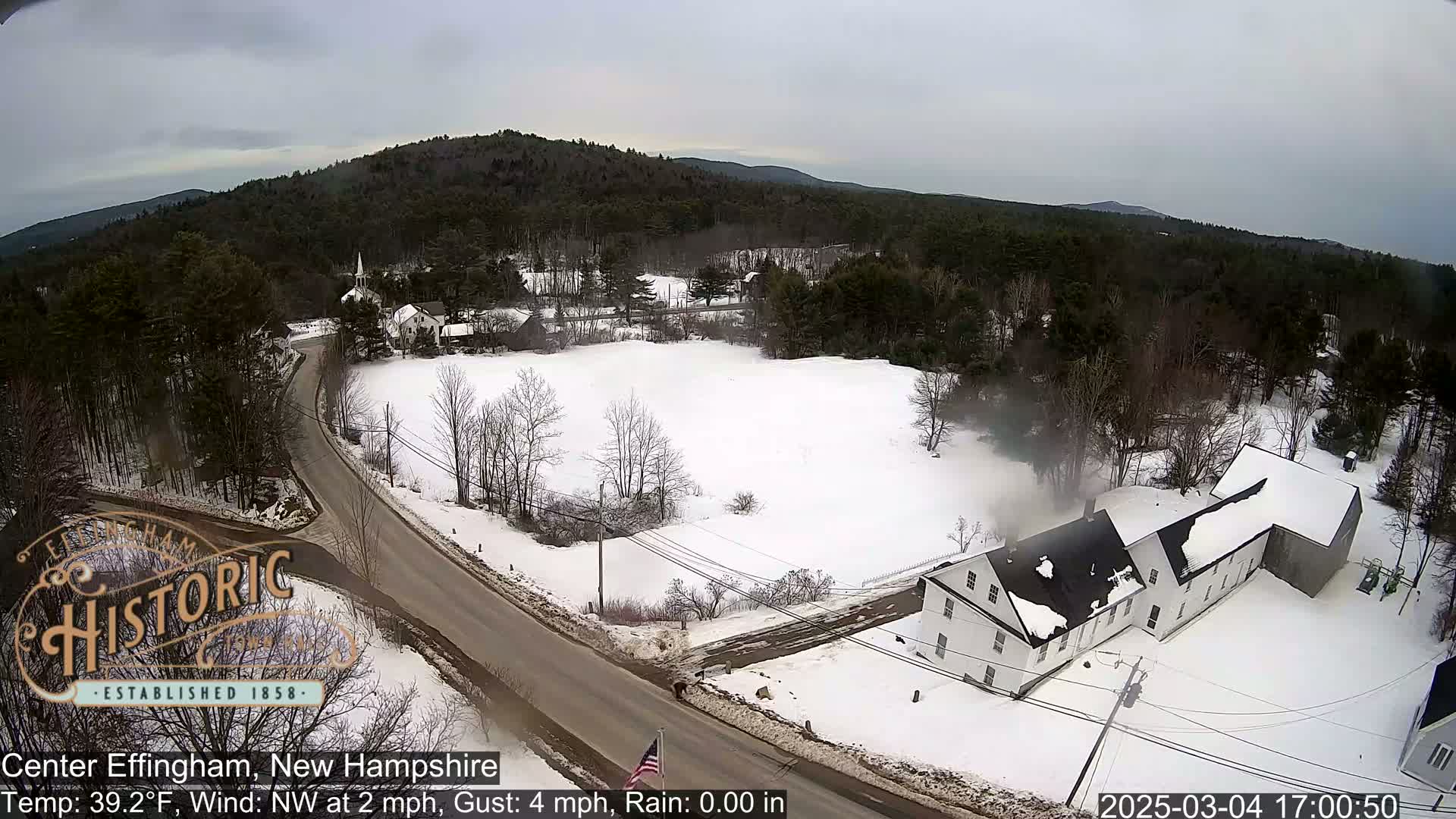 A snow-covered town with a road and buildings is viewed from above on a cloudy day.