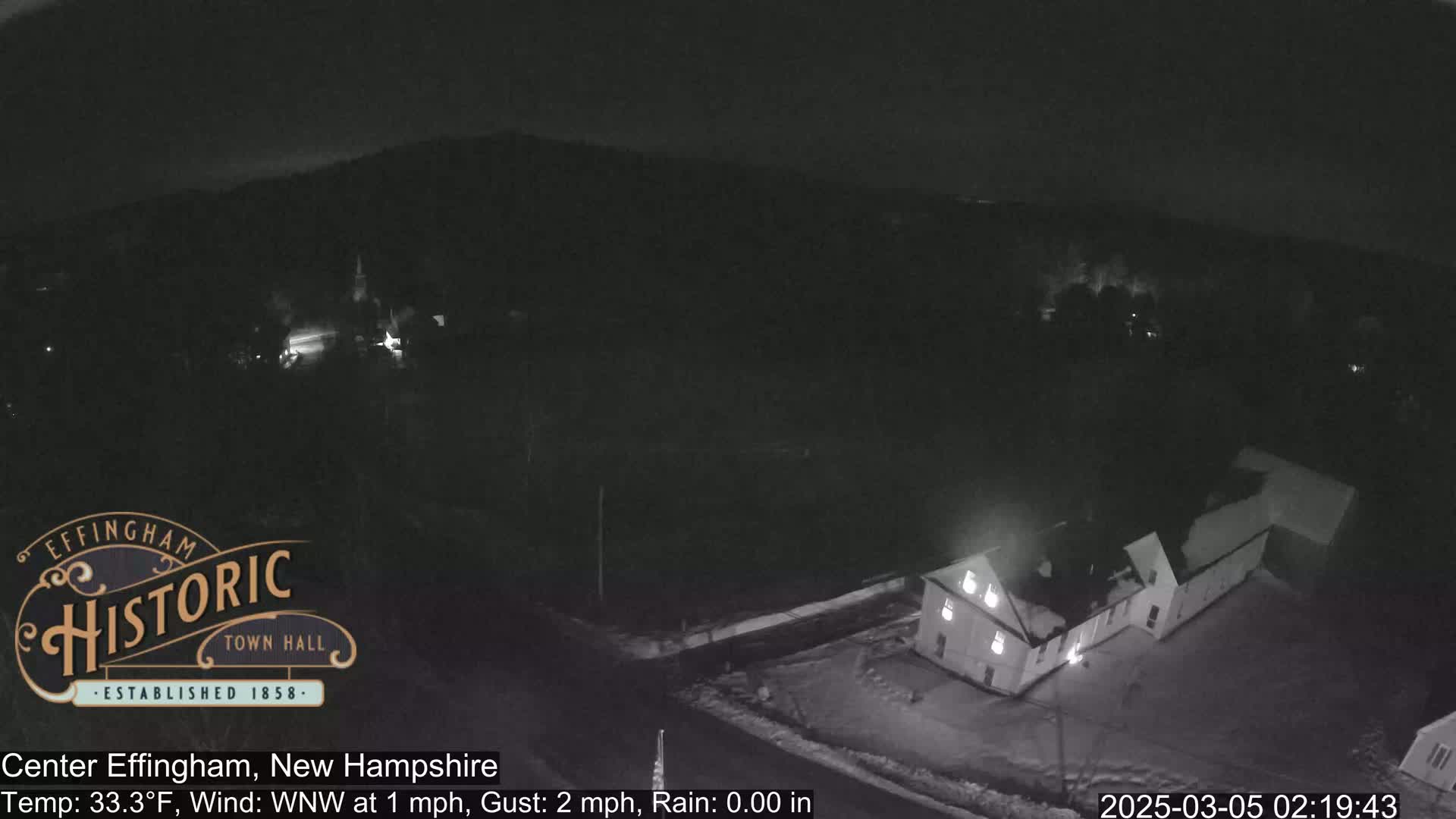 A nighttime, low-light aerial view of a building complex and surrounding snow-covered ground under a clear sky with a temperature of 33.3°F, a 1 mph wind from the west-northwest, and no rain.