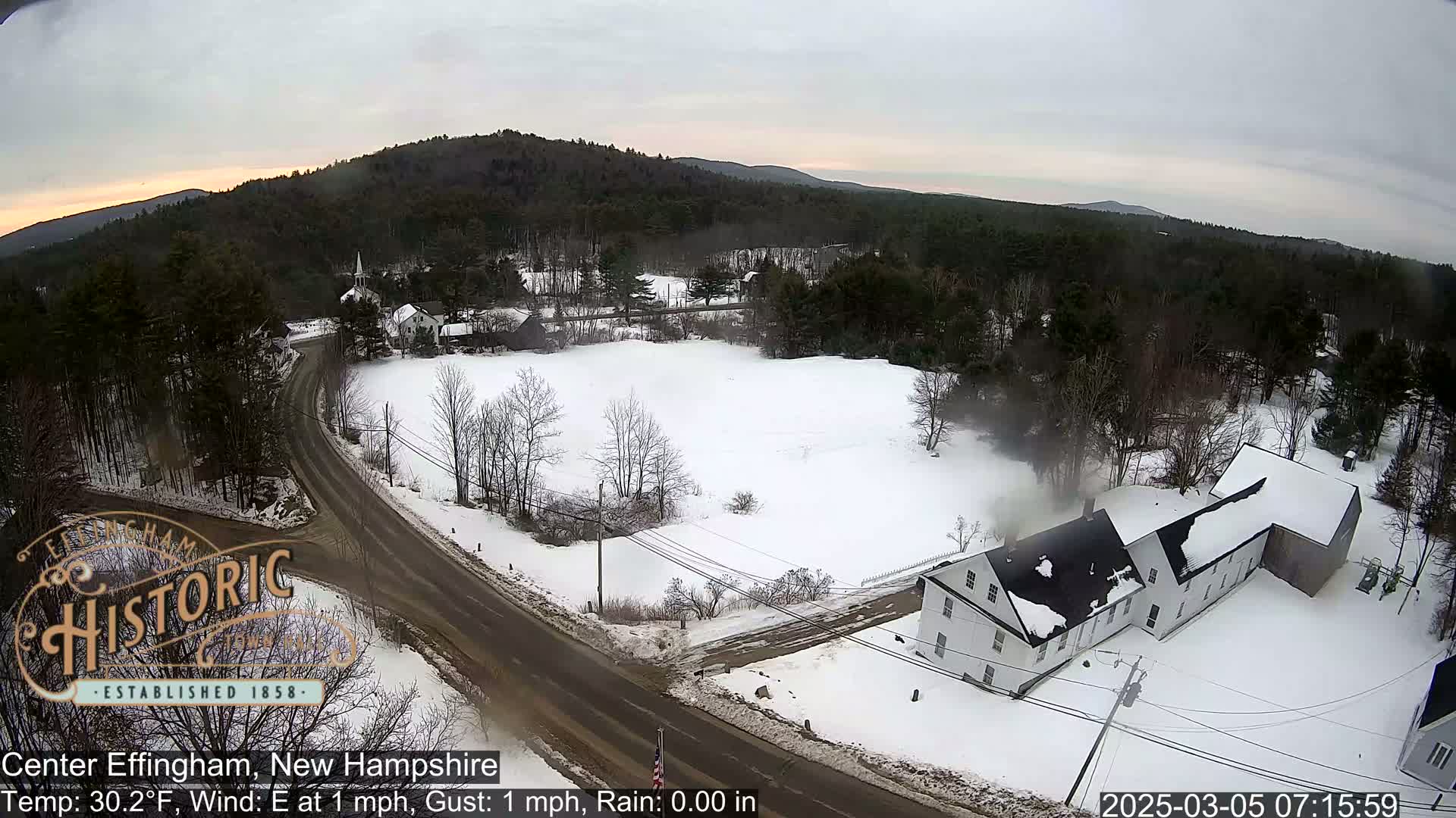 A snow-covered landscape with a road and buildings is viewed from above under a cloudy sky.