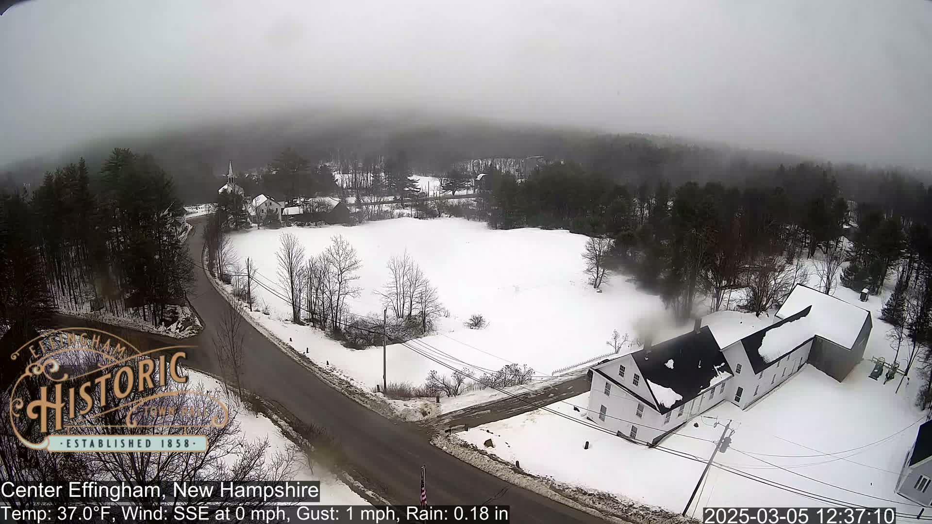 A snow-covered landscape with buildings and a road, viewed on an overcast day with light rain.
