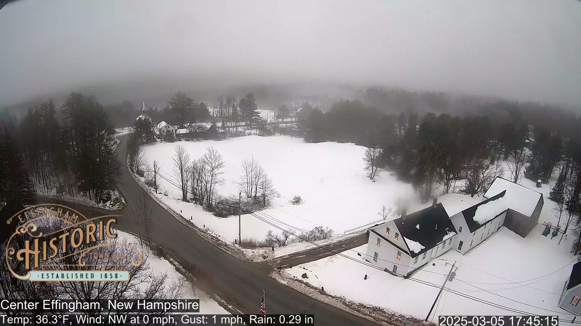 A snow-covered landscape with buildings and a road is visible under foggy conditions.