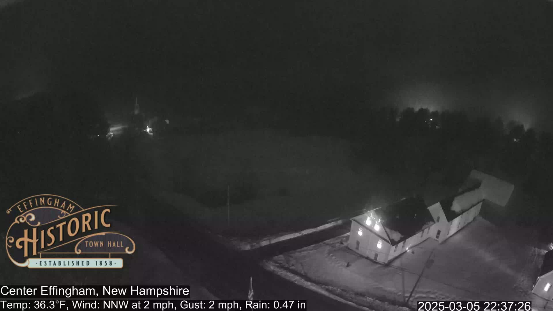 A nighttime, snowy scene shows a building and road under a dark sky with light rain.