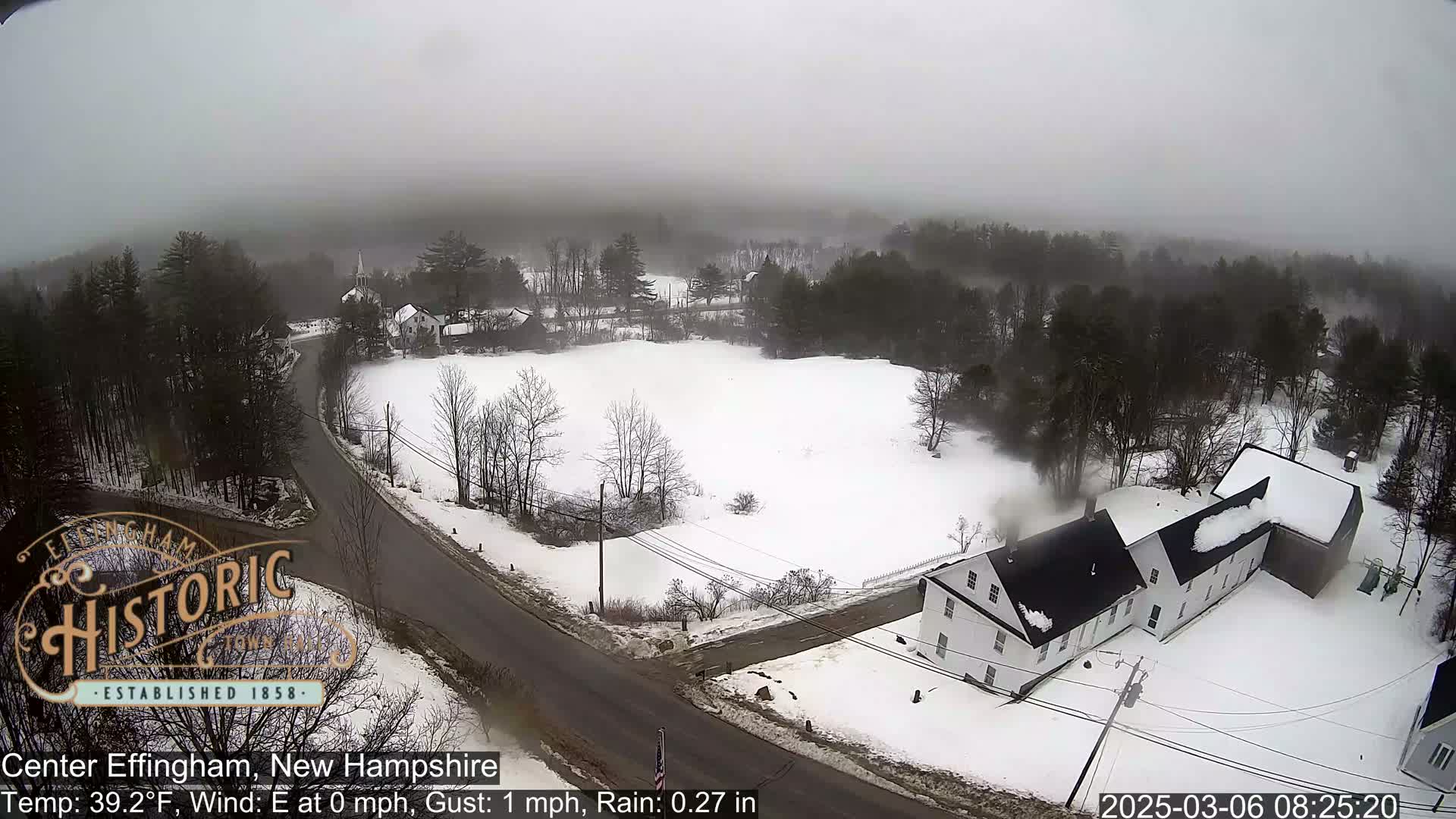 A foggy, snowy, rural landscape with a road and several buildings, including a long, white house, is viewed from above.