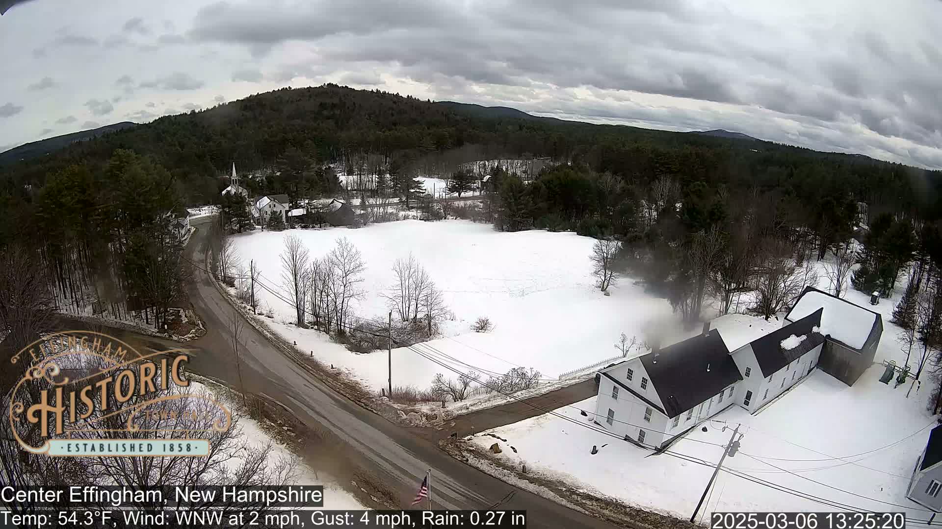 A snow-covered landscape with houses and a road, under a cloudy sky.