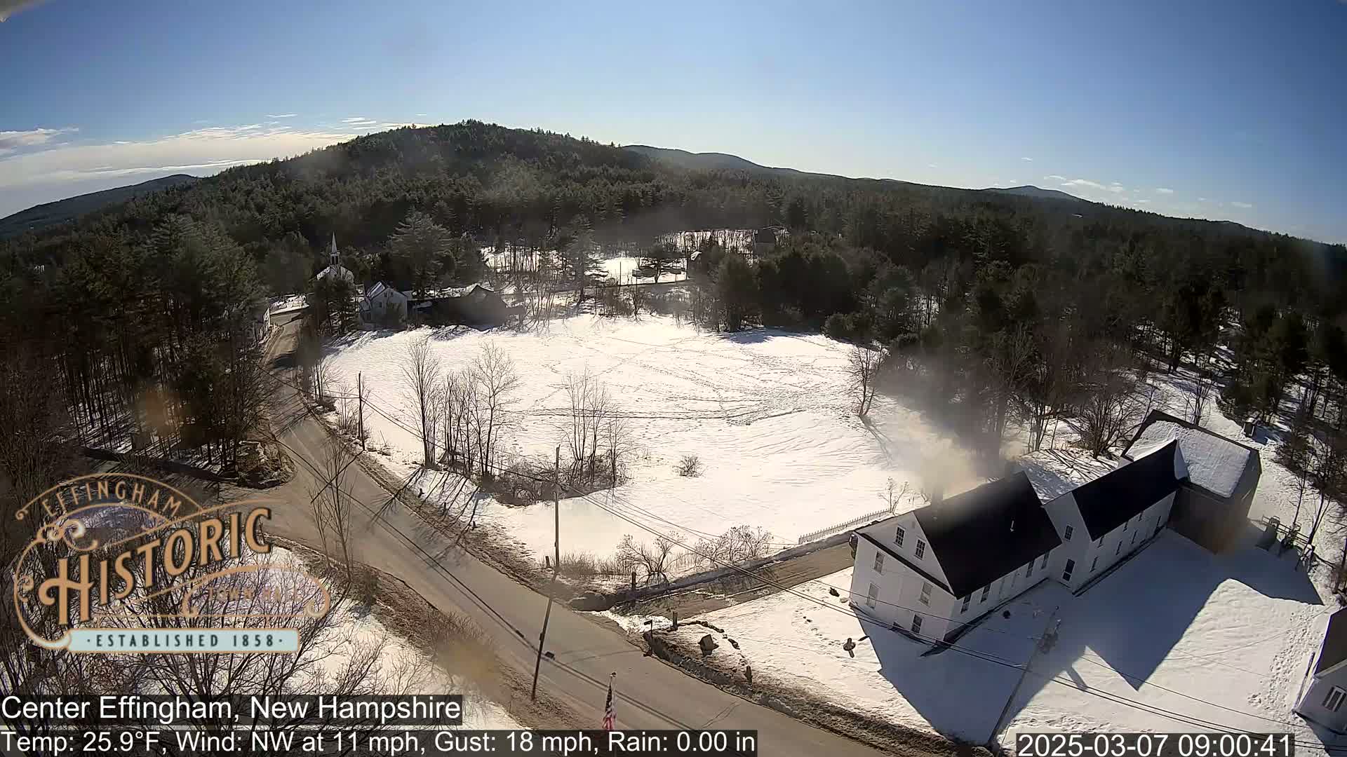 A snow-covered village scene is viewed from above on a sunny day, with a northwest wind and some bare trees.