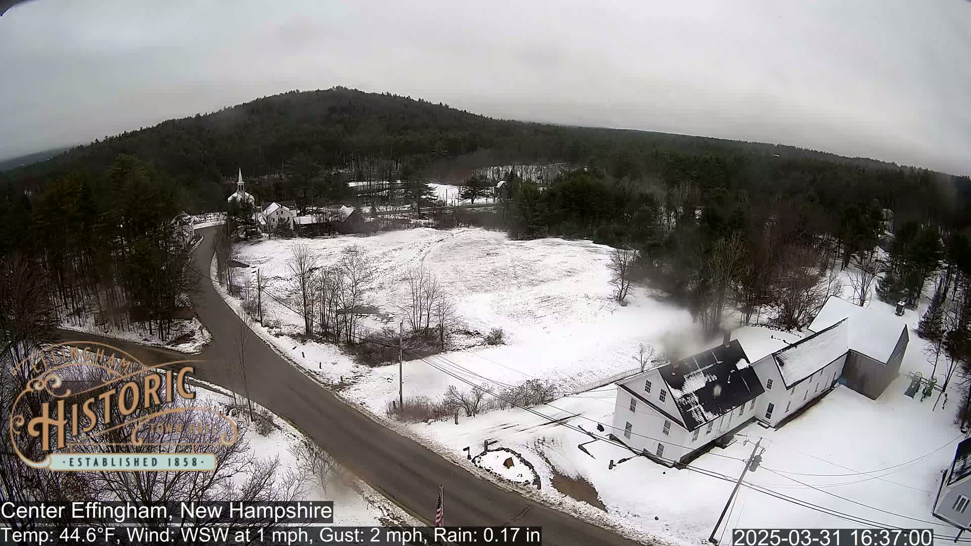 An aerial view of a snow-covered town with a road, buildings, and trees under a cloudy sky with light rain.