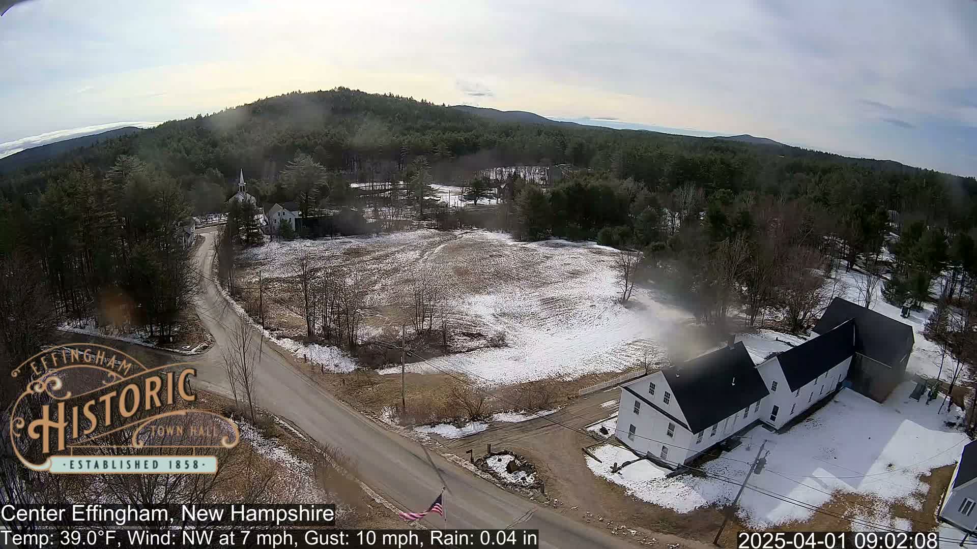 An aerial view shows a small town with a road, some buildings, and patchy snow cover on the ground under a cloudy sky.