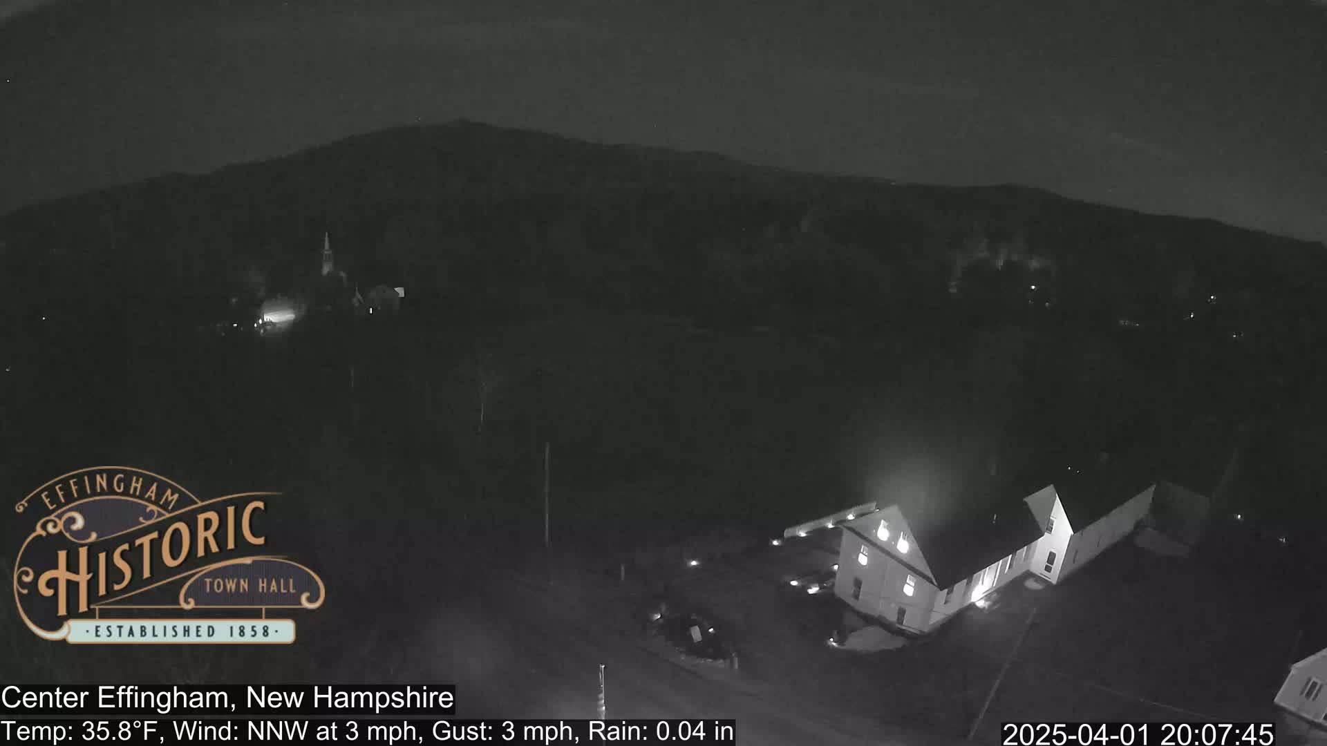A nighttime, rain-affected view shows a building, possibly a town hall, and surrounding landscape under a dark sky.