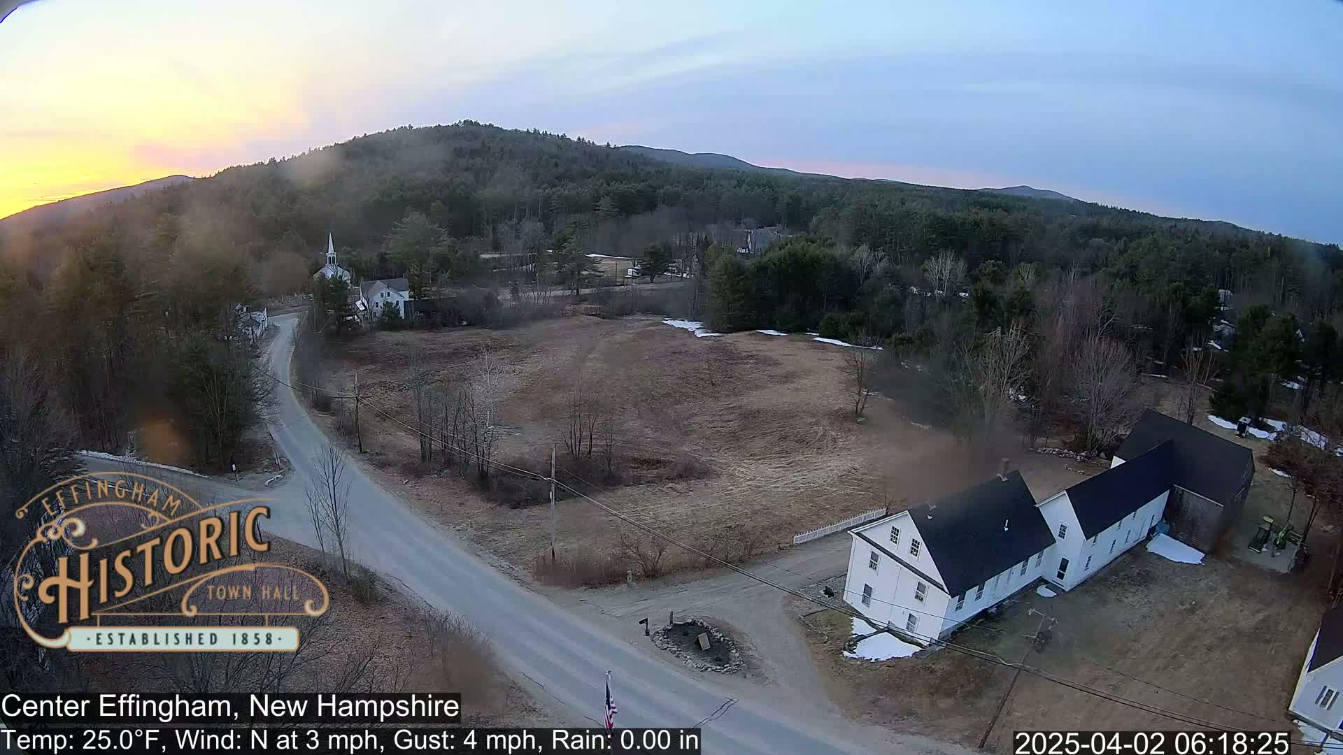 An aerial view shows a rural town at sunrise with a light dusting of snow on the ground and a mostly clear sky.