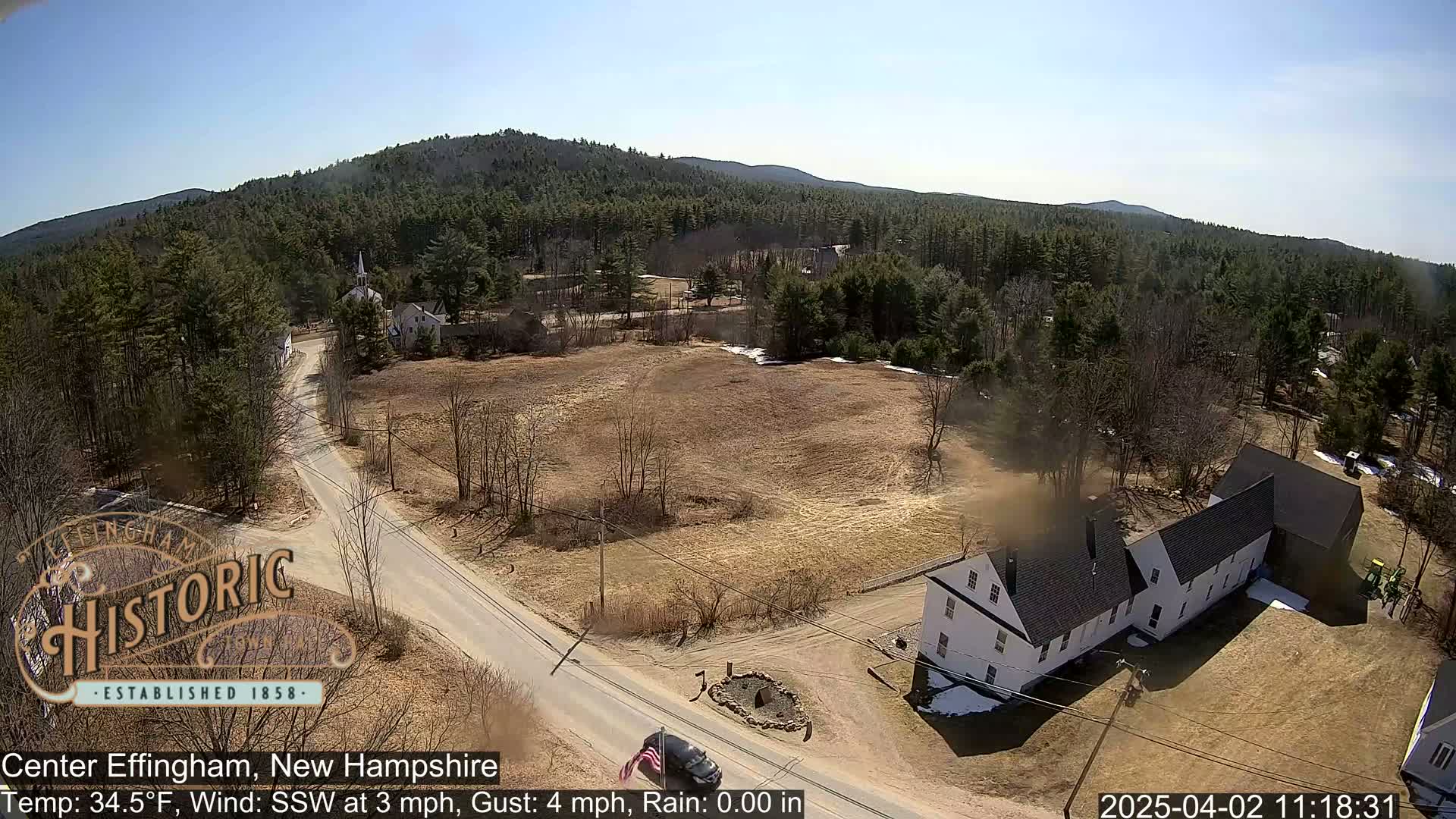 A high-angle view of a small town nestled in a valley of mostly bare trees under a clear blue sky, with a long, white building near a road intersection.