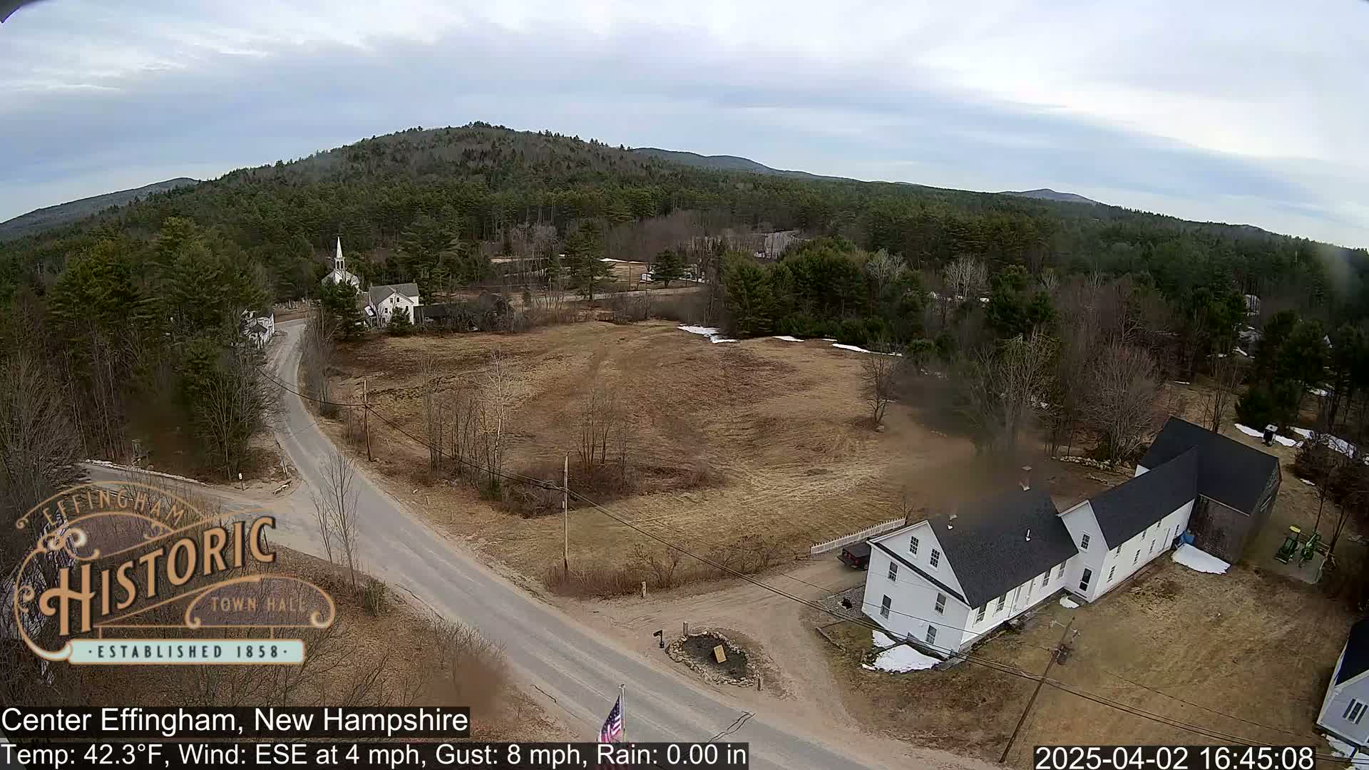 An aerial view shows a rural town with a church, houses, and roads under partly cloudy skies.