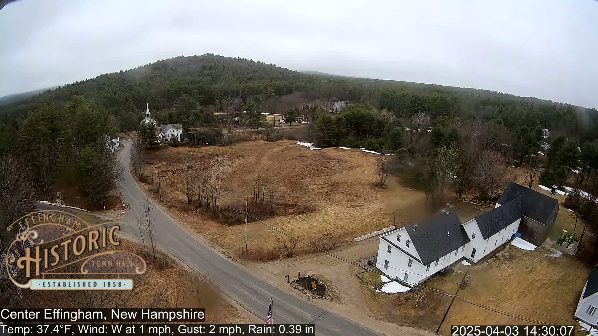 An aerial view shows a small town nestled in a wooded area on an overcast day with light rain.