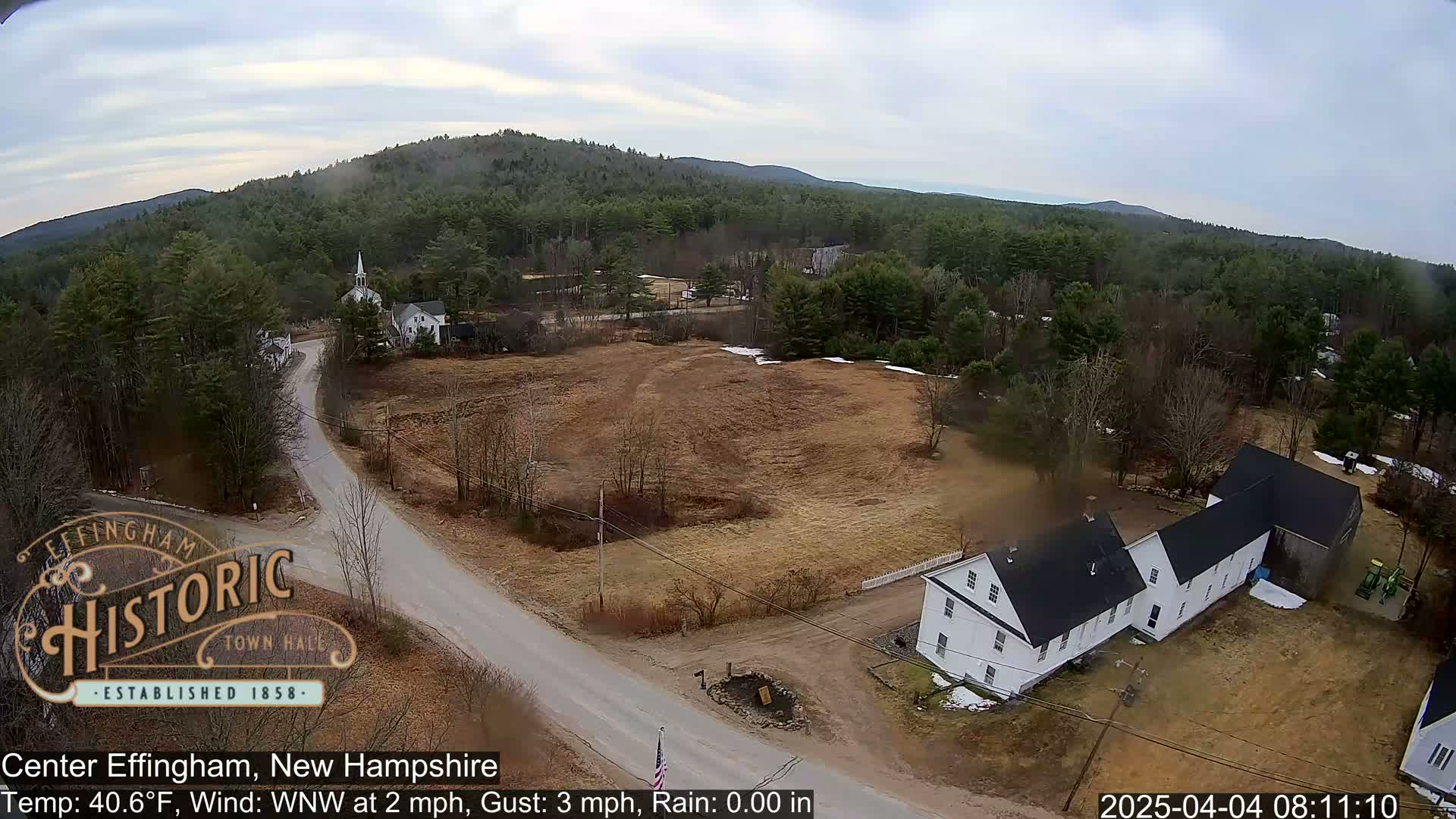 An aerial view shows a rural town with a road, buildings, and a mostly brown, sparsely vegetated landscape under a partly cloudy sky.
