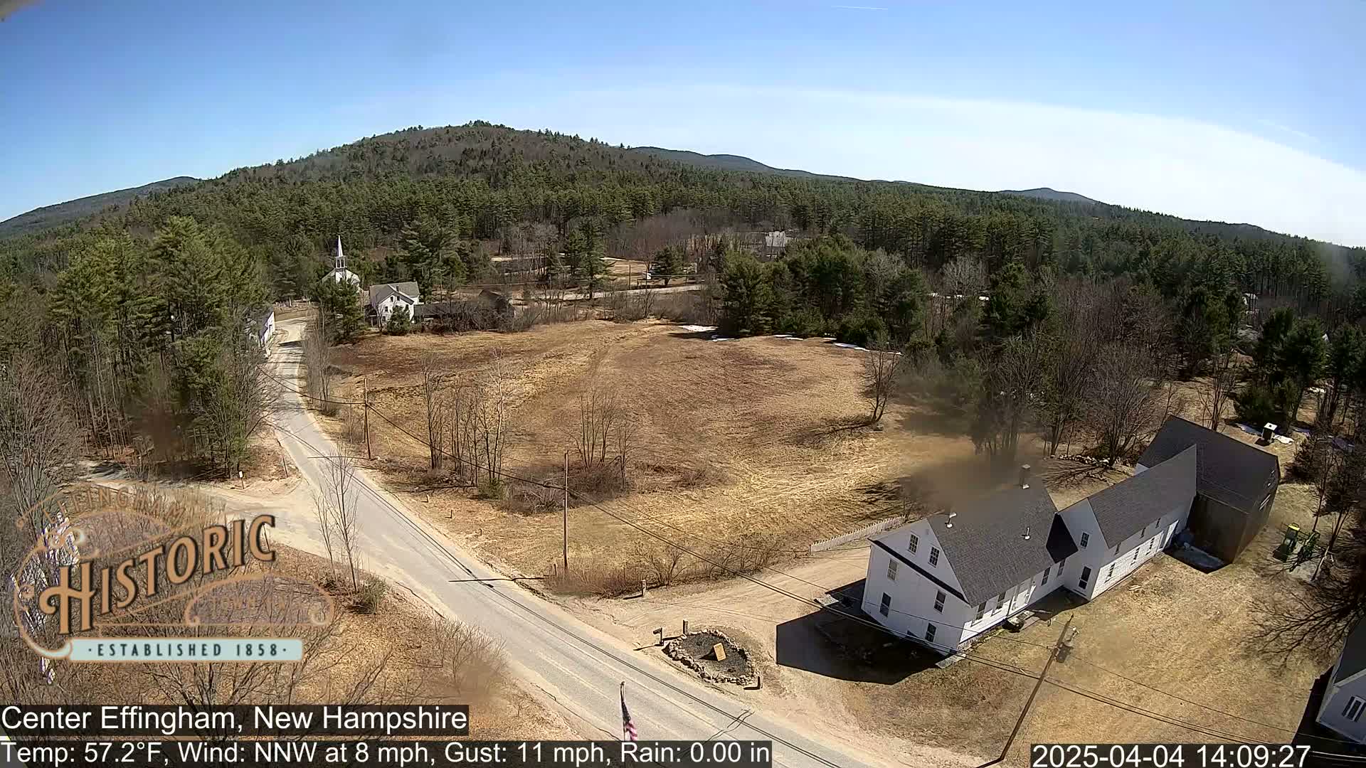 A high-angle view of a small town nestled among a forest of evergreens under a clear blue sky, with light rain and a northwest wind.