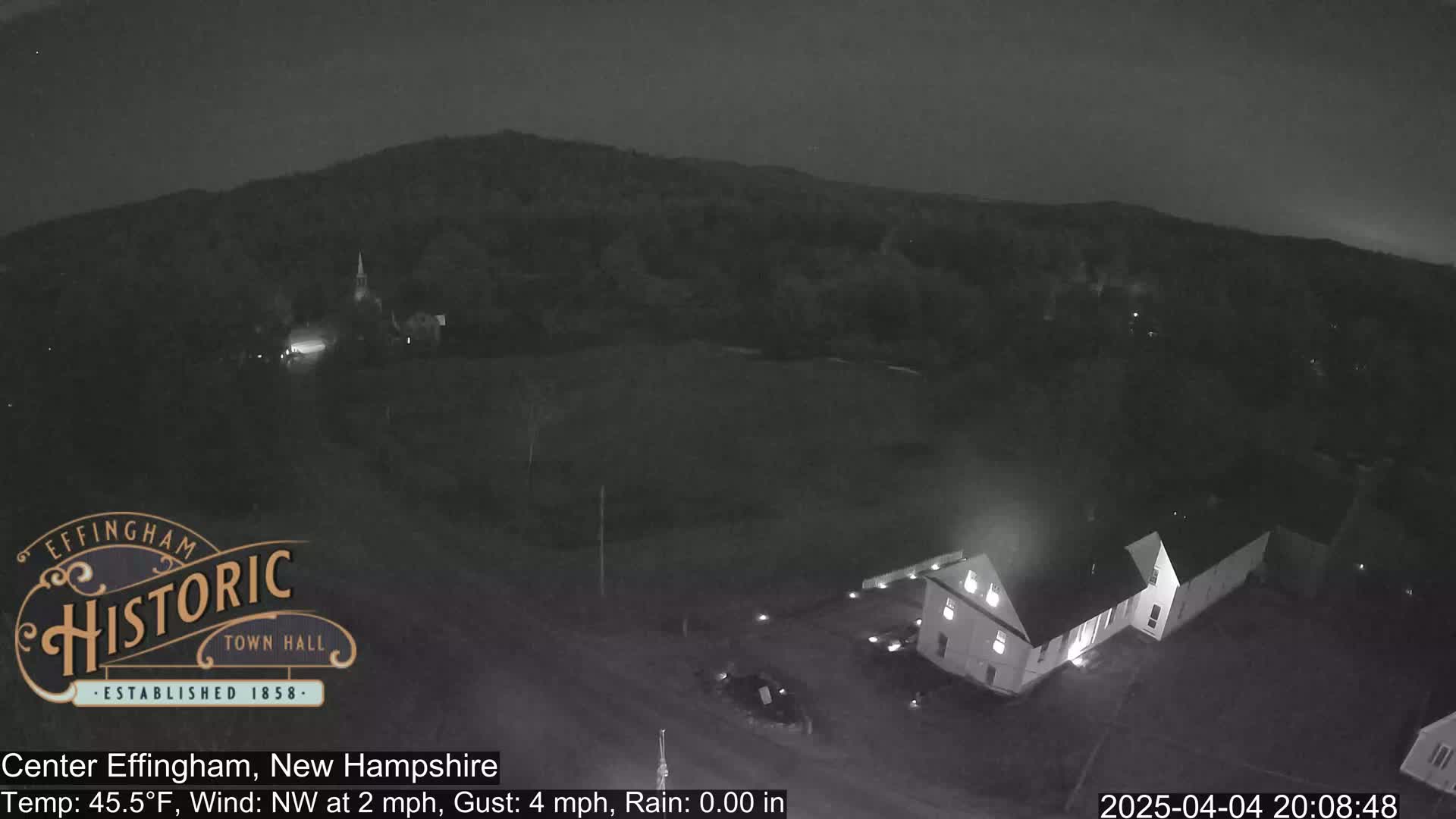 A nighttime, low-light view of a small town nestled among hills, with a northwest wind at 2 mph and gusts up to 4 mph.
