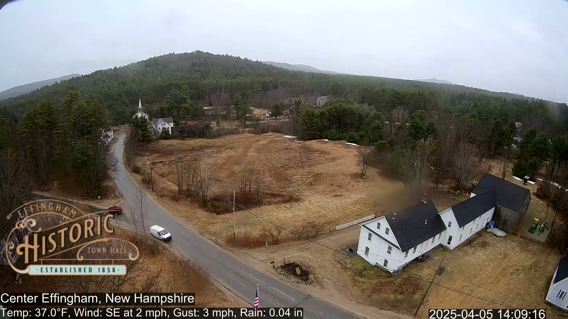 A lightly raining, elevated view of a small town with a church steeple, houses, and vehicles on a road, surrounded by brown fields and a forested hillside.