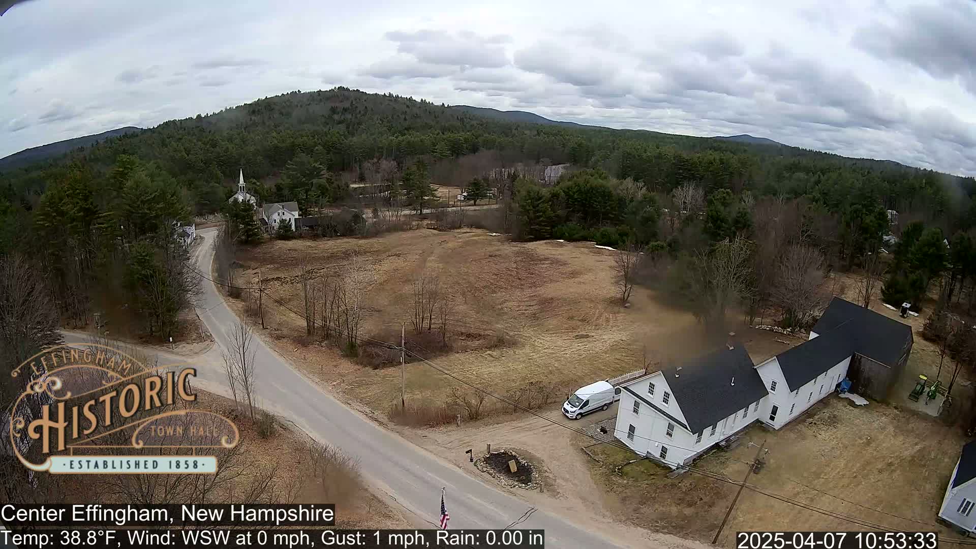 An aerial view shows a rural town with a church steeple and several houses under a cloudy sky.