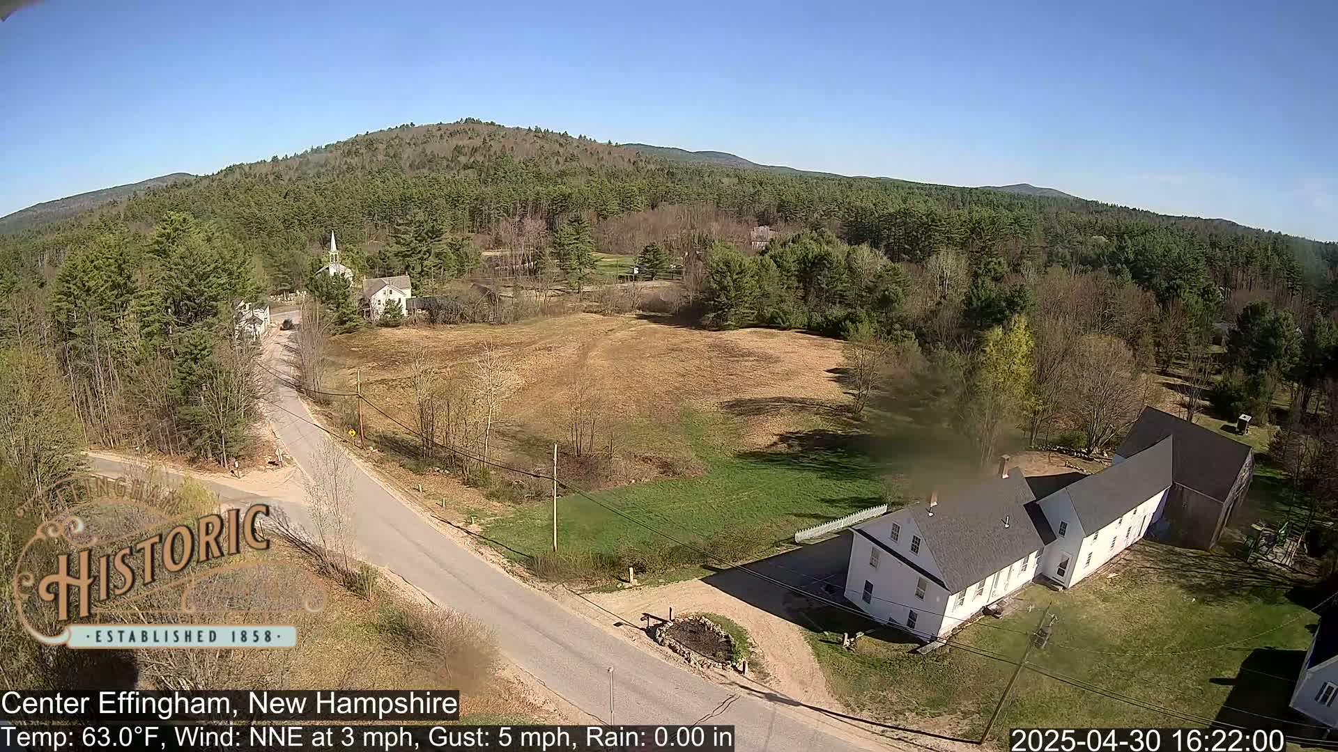 A sunny, rural landscape with a church steeple, houses, and a road intersection, nestled among green trees and hills.