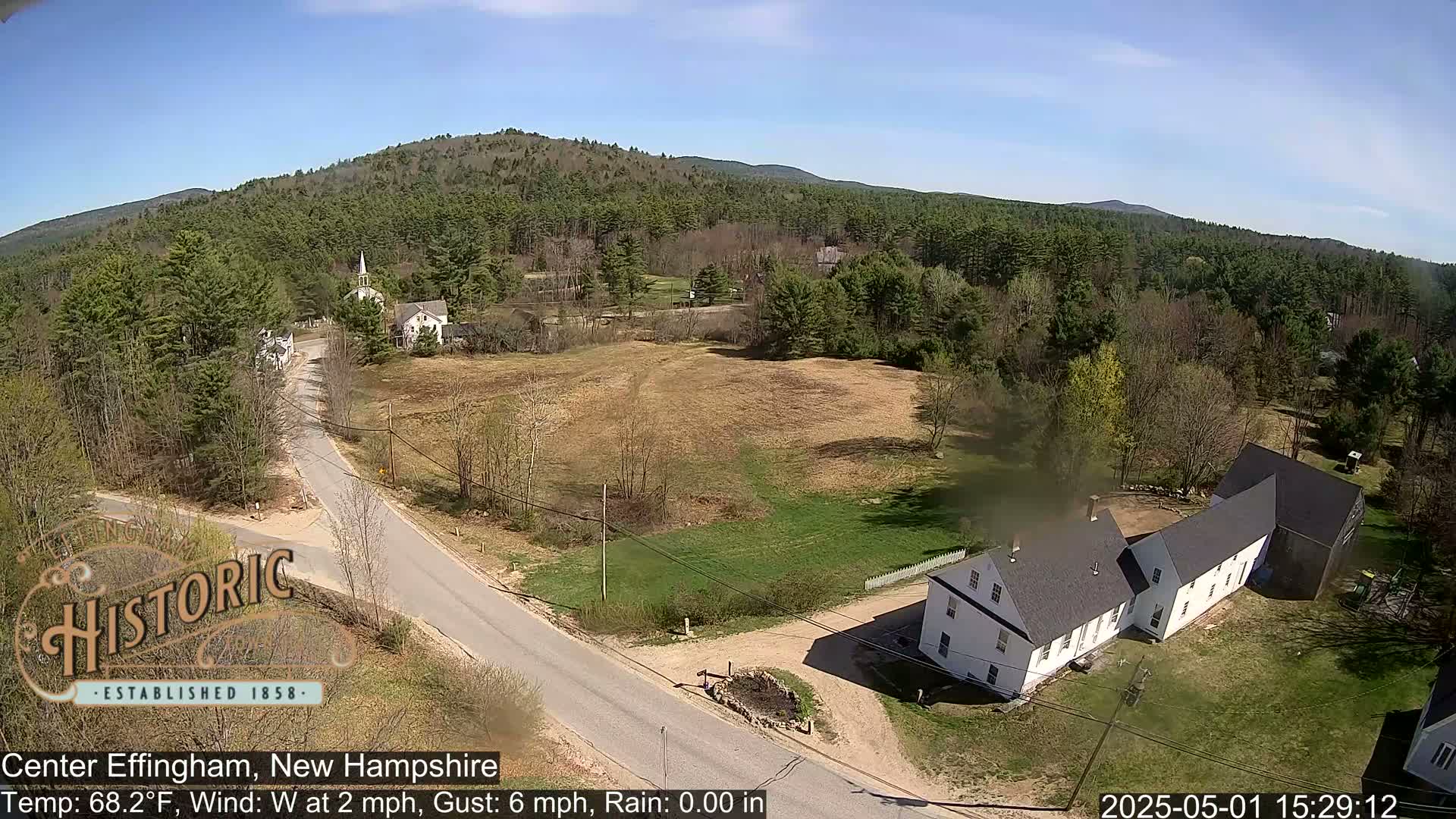 A high-angle view of a rural town on a sunny day with a light westerly wind shows a road intersecting near several buildings nestled amongst trees and open fields, with mountains in the distance.