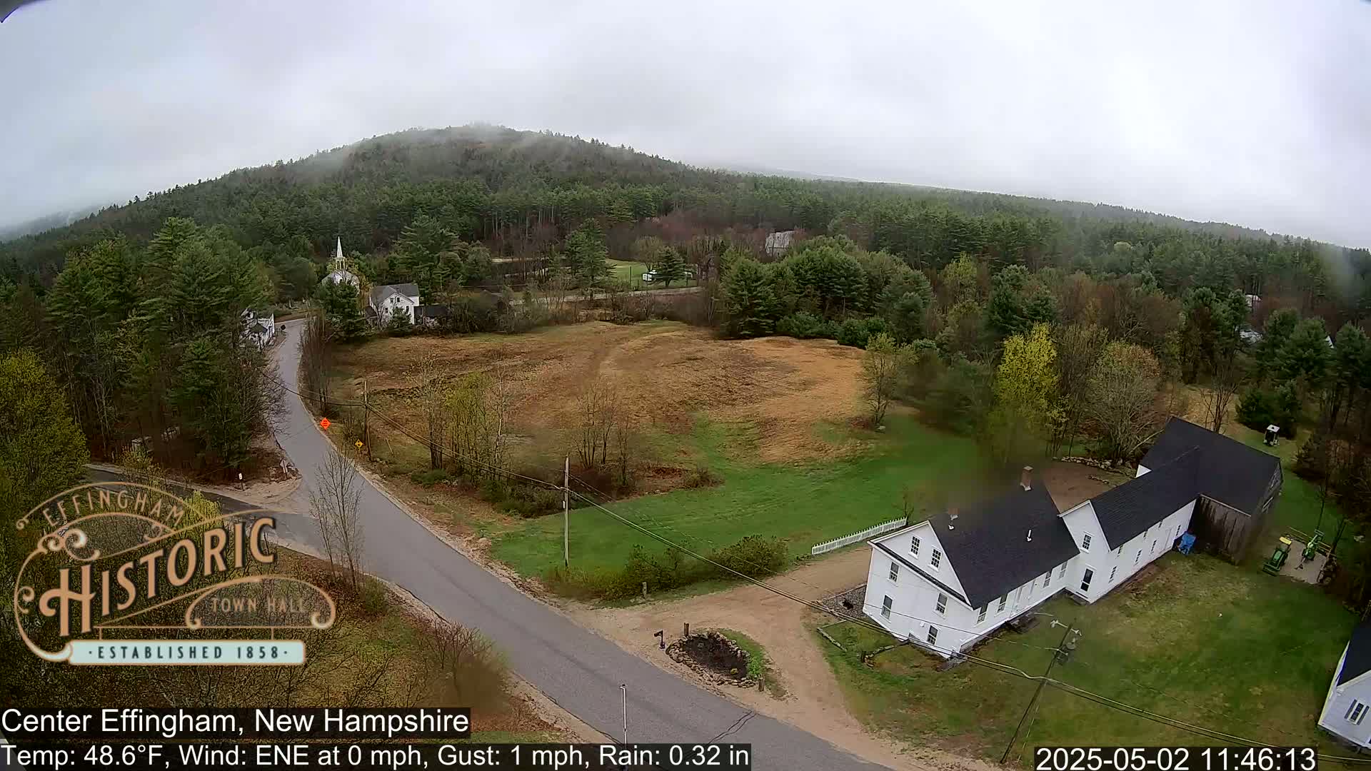 A rainy, overcast view of a small town nestled among green hills and forests, showing a road intersecting near a cluster of white houses.